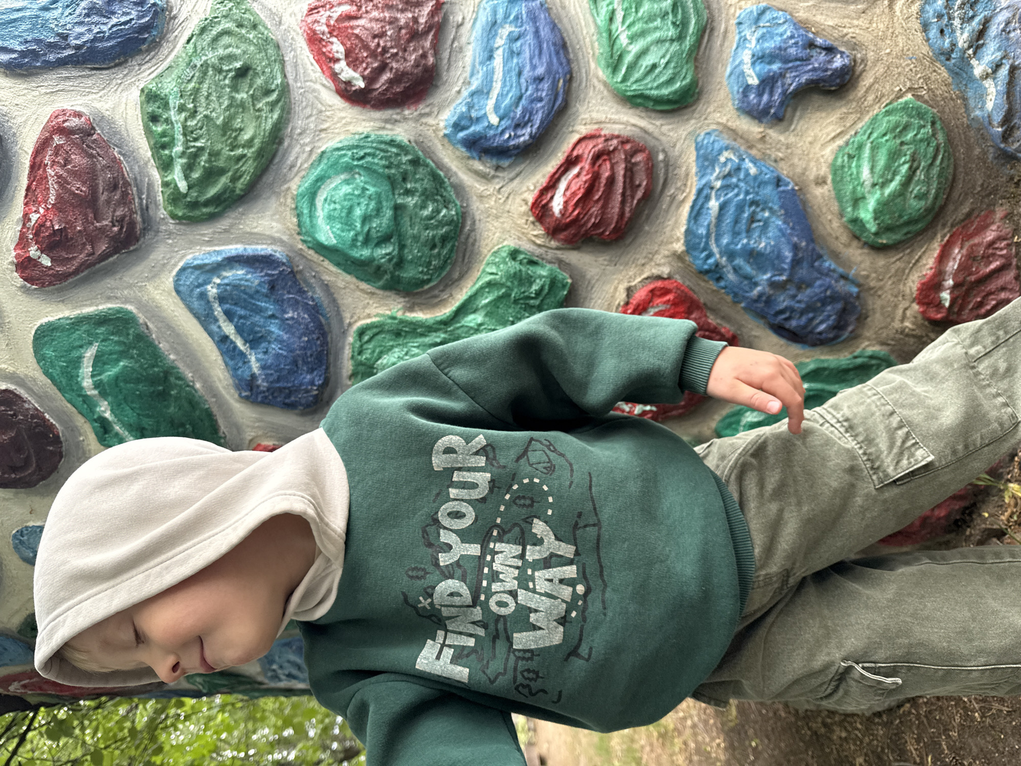 A child wearing a green hooded sweatshirt with the phrase "Find Your Own Way" and gray cargo pants is sitting next to a wall with colorful, textured stone patterns of red, blue, and green. The scene appears to be outdoors, possibly in a park or playground. The location may be Chaikovsky, Russia, but there is no additional information in the image to confirm the location.