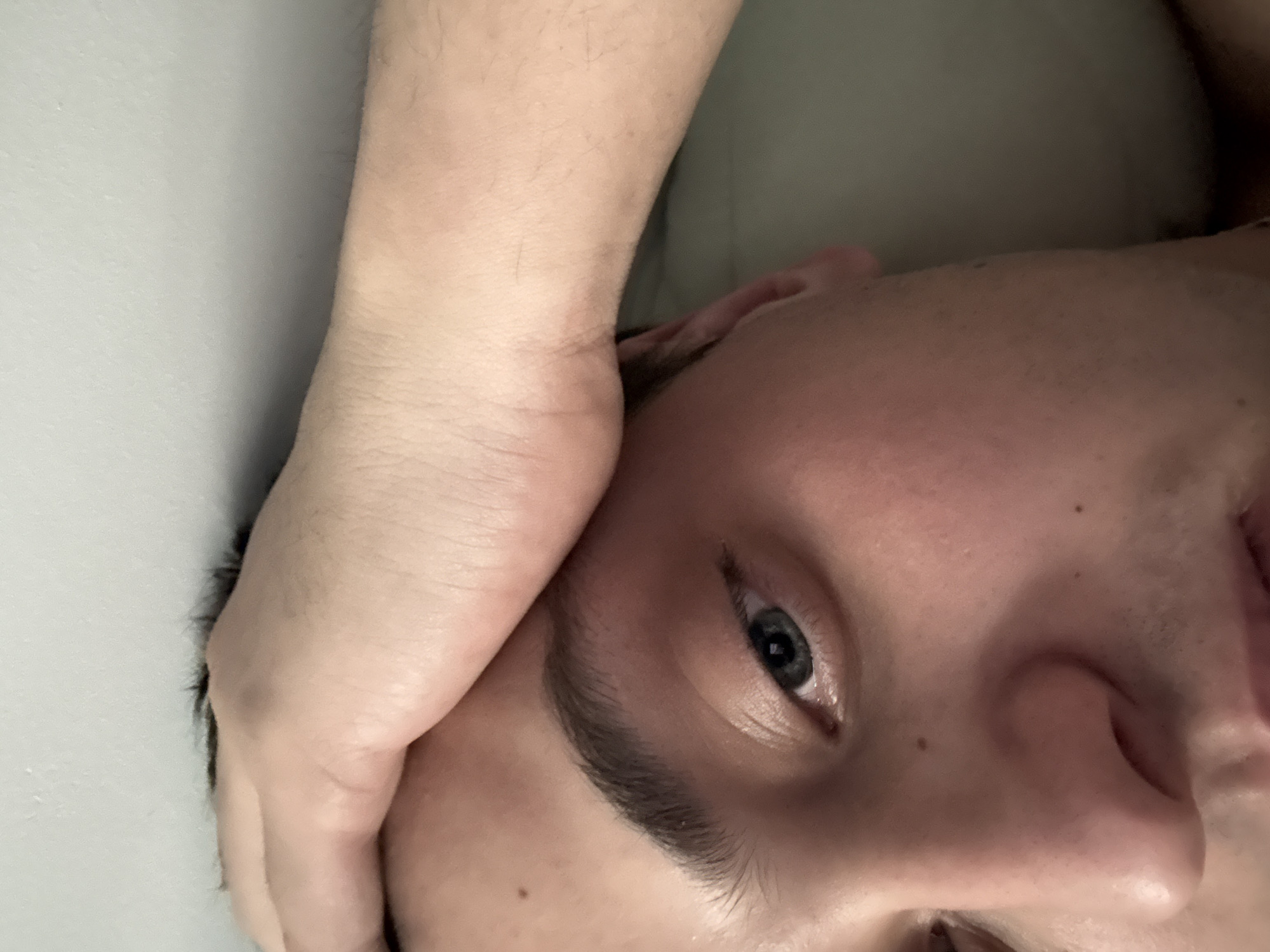 Close-up shot of a person's face, partially obscured by their hand. The individual has fair skin, dark eyebrows, and a visible eye with a gray iris. The person's hand is positioned near their forehead. The background is out of focus and shows a wall and possible bedding. The image does not provide sufficient information to determine the location as Vidnoye, Russia, or to ascertain specific actions or interactions.