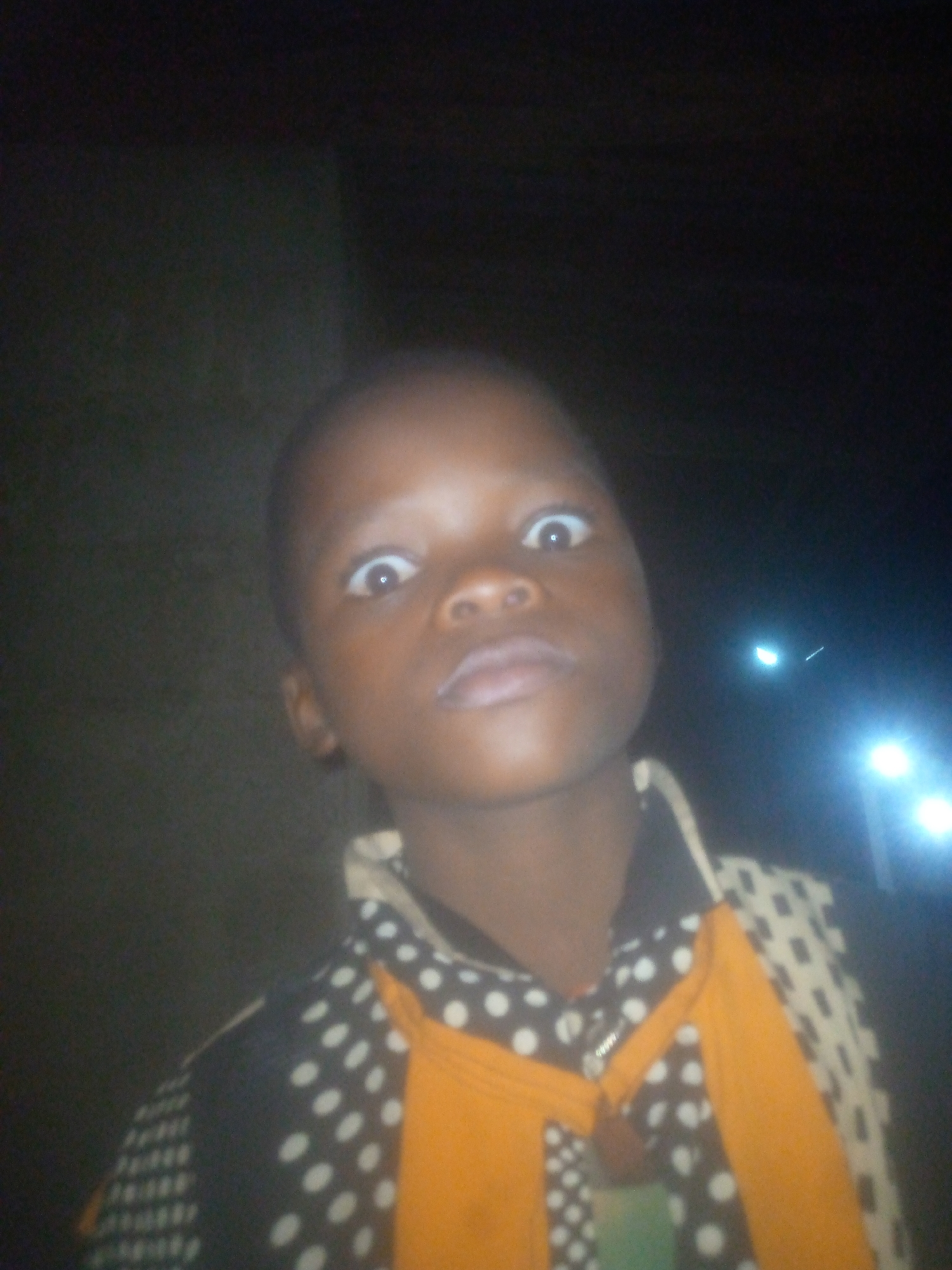 A young person with dark skin stares wide-eyed at the camera. The subject is wearing a black and white polka-dot shirt with yellow bands on the front. The photo was likely taken at night, with two bright lights visible in the background. The location is unknown, Unknown. A wall is visible to the left of the subject.