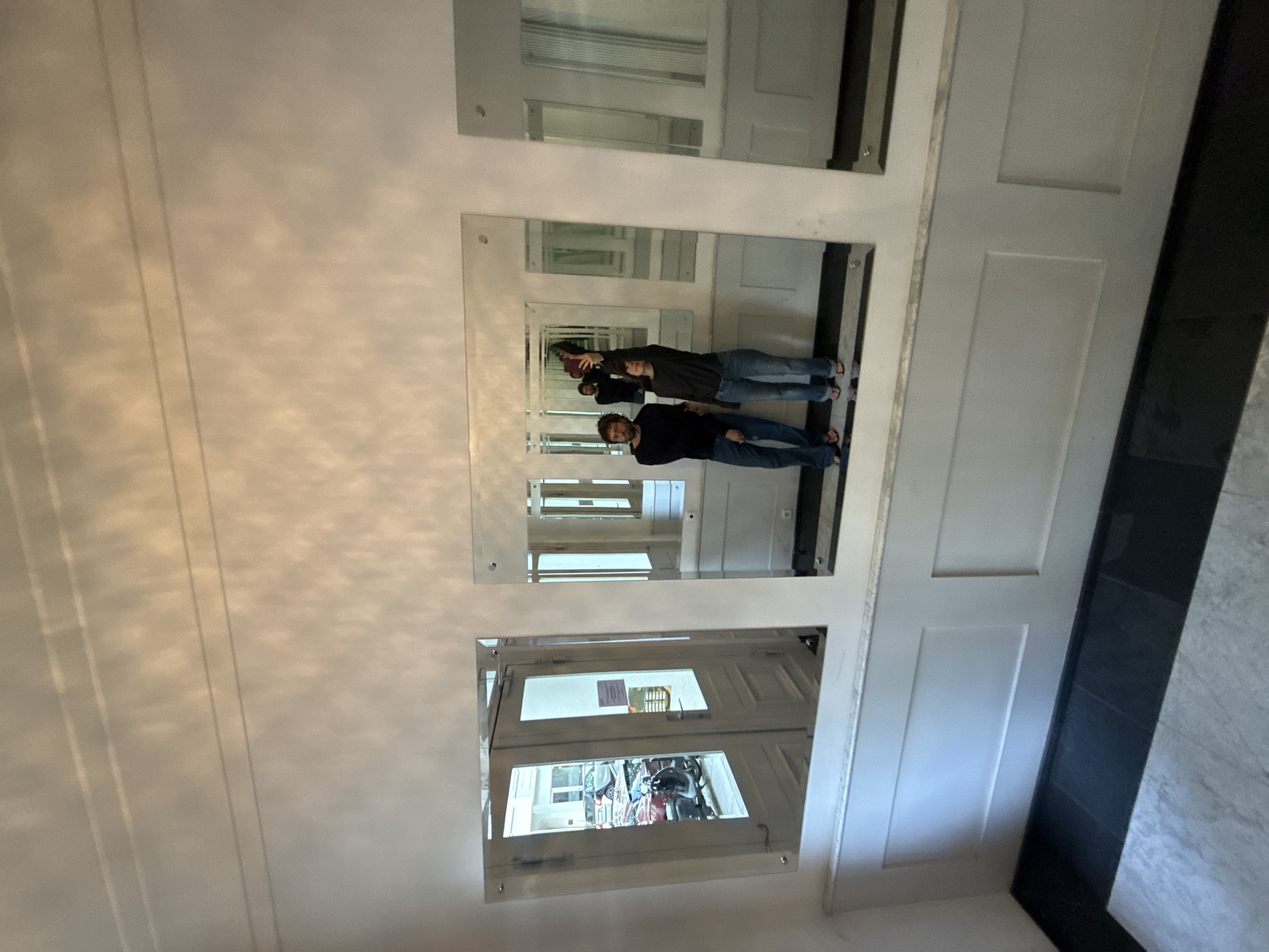 Two men, reflected in a series of mirrors, appear in a hallway. One man is taking a picture with his phone. The hallway contains multiple doors, and a rack of bicycles is visible through one door. The location is likely Berlin, Germany.