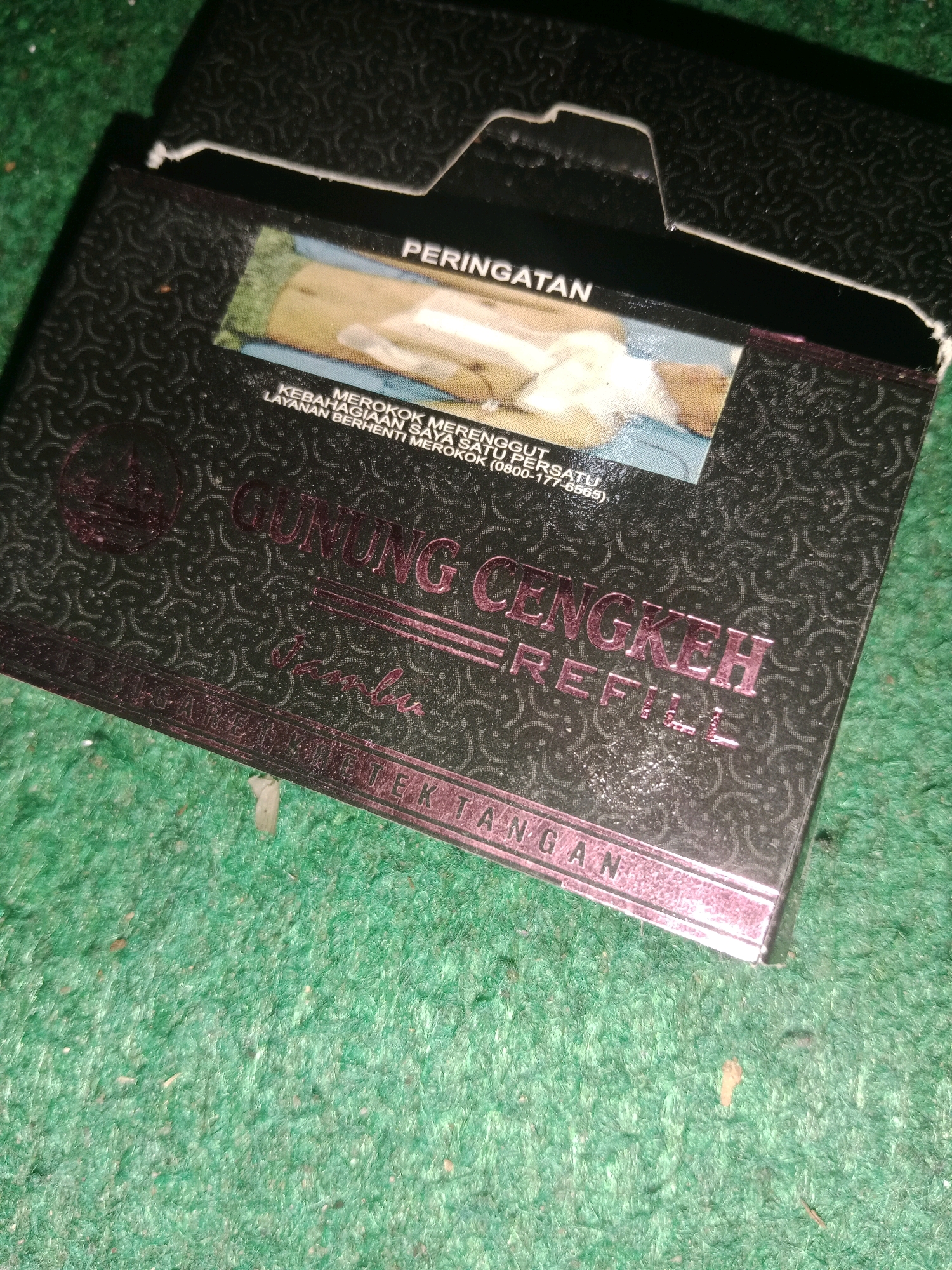 A close-up, high-angle shot shows a pack of "GUNUNG CENGKEH REFILL" cigarettes on a green, textured surface, likely carpet. The pack is dark with a repeating pattern and has a warning label in Indonesian that reads "PERINGATAN MEROKOK MERENGGUT KEBAHAGIAAN SAYA SATU PERSATU LAYANAN BERHENTI MEROKOK (0800-177-6565)" which translates to "WARNING SMOKING SNATCHES MY HAPPINESS ONE BY ONE QUIT SMOKING SERVICE". The pack is partially open, revealing a small image of what appears to be a person smoking. The setting is likely indoors, and given the product, it could be found in many locations in Ringinrejo, Indonesia, where such cigarettes are sold. There are no people visible in the image.