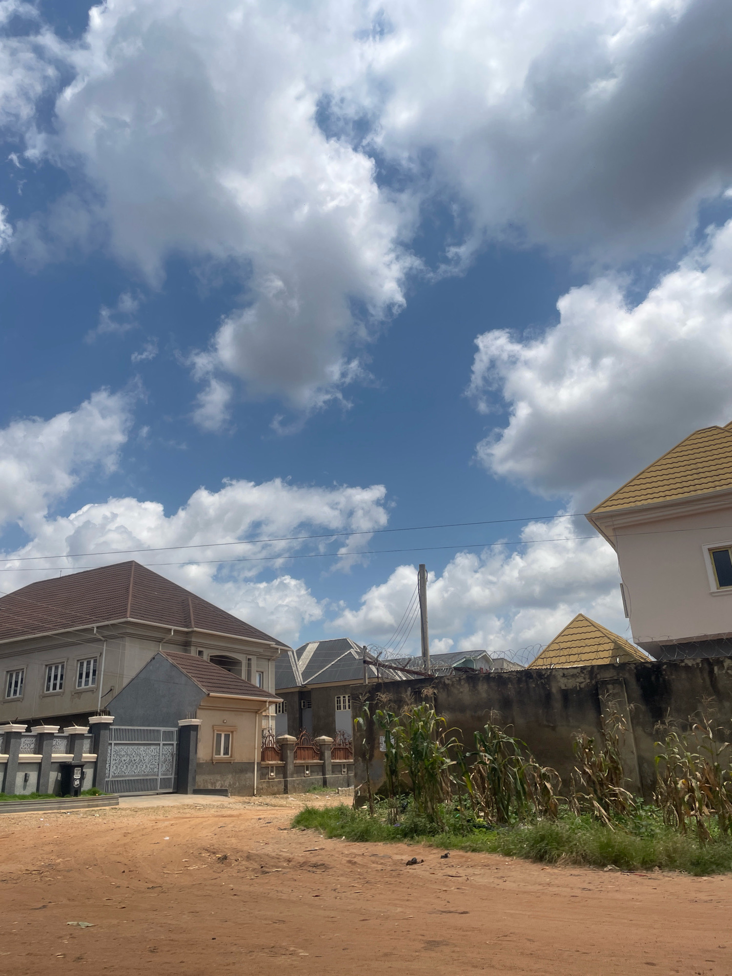This is a daytime, eye-level, outdoor, medium-shot photograph taken in Abuja, Nigeria, likely on an unpaved, dirt road, indicating a developing or less urbanized area of the city. The scene depicts several modern, two-story residential buildings with distinct architectural styles, some featuring brown and yellow tiled roofs. A tall concrete wall, partially overgrown with dry corn stalks and vegetation, runs horizontally across the middle of the frame, separating the road from the properties. The sky above is a vibrant blue, dotted with scattered, fluffy white clouds, suggesting fair weather. In the foreground, the reddish-brown dirt road occupies the lower portion of the image. The composition is dominated by the architecture and the sky, with the wall and vegetation serving as a mid-ground element. There are no people visible in the frame. The overall impression is one of a quiet residential street in a sunny, possibly hot, climate.