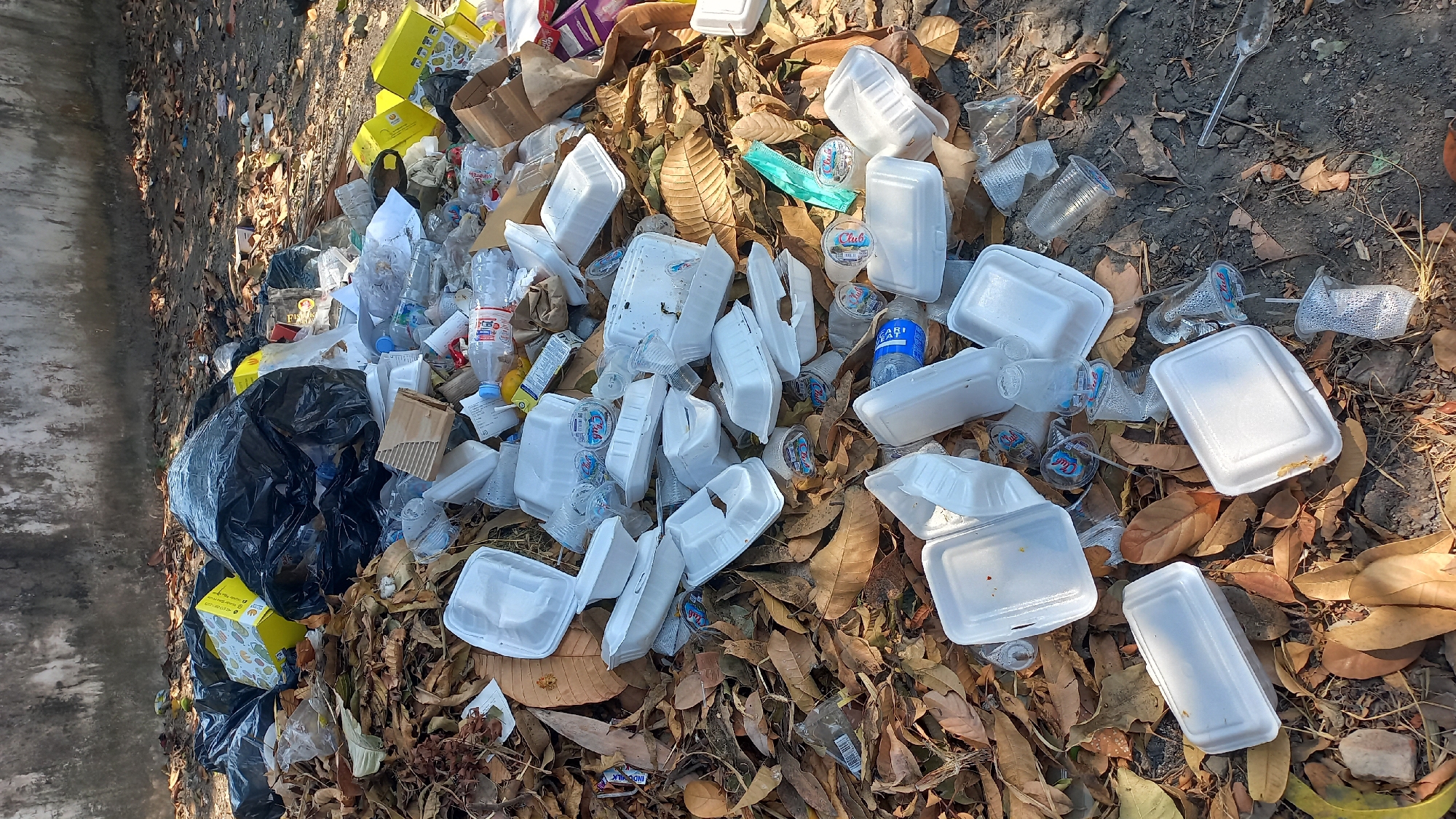The image depicts a large pile of trash in an outdoor urban setting, likely located in Kayen Kidul, Indonesia. The refuse consists of black garbage bags, various discarded food containers, primarily white styrofoam clamshells and boxes, plastic bottles, and a significant amount of fallen leaves and debris. The trash is spread across a dirt and gravel ground surface, with a weathered, concrete wall forming the background. The scene appears to be a neglected area, possibly a roadside or a less-trafficked alleyway, characterized by the accumulation of waste. Sunlight casts shadows, suggesting it is daytime. No people are visible in the image.