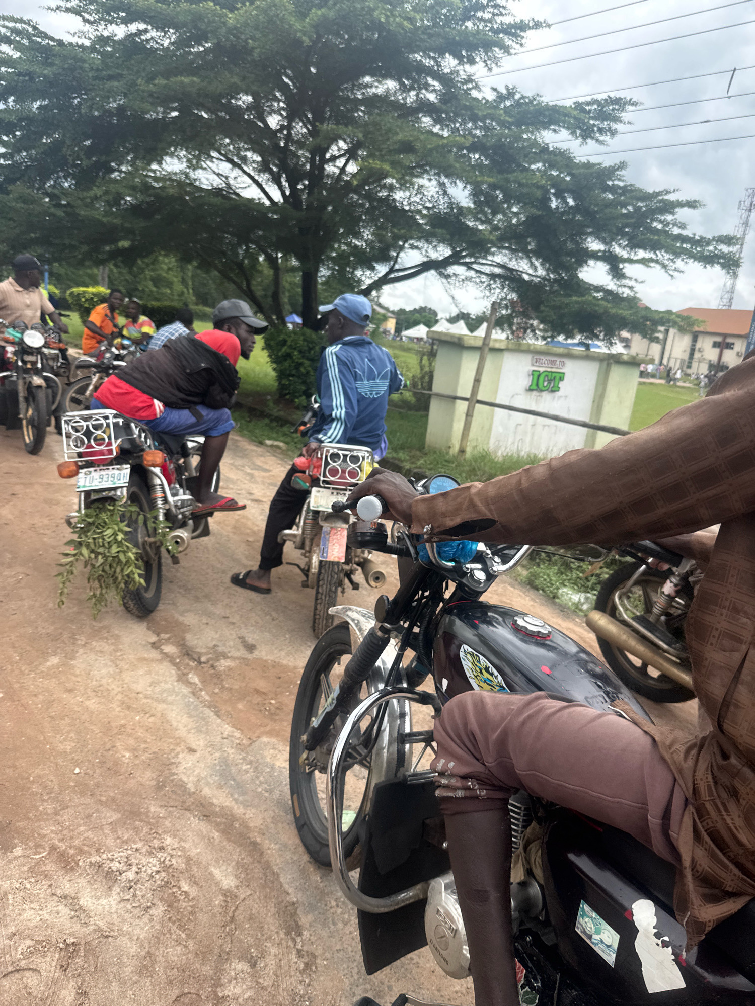 In Ihiagwa, Nigeria, a group of men on motorcycles are gathered on a dirt road. The scene is dominated by the motorcycles, with several individuals seated on them. One man in a red shirt and black jacket is prominently featured, his motorcycle adorned with a bunch of green leaves. Another man in a blue tracksuit is also visible on his motorcycle. In the background, a building with "WELCOME TO ICT" signage is present, along with trees and a cloudy sky. The overall impression is a snapshot of daily life and transportation in a Nigerian city.