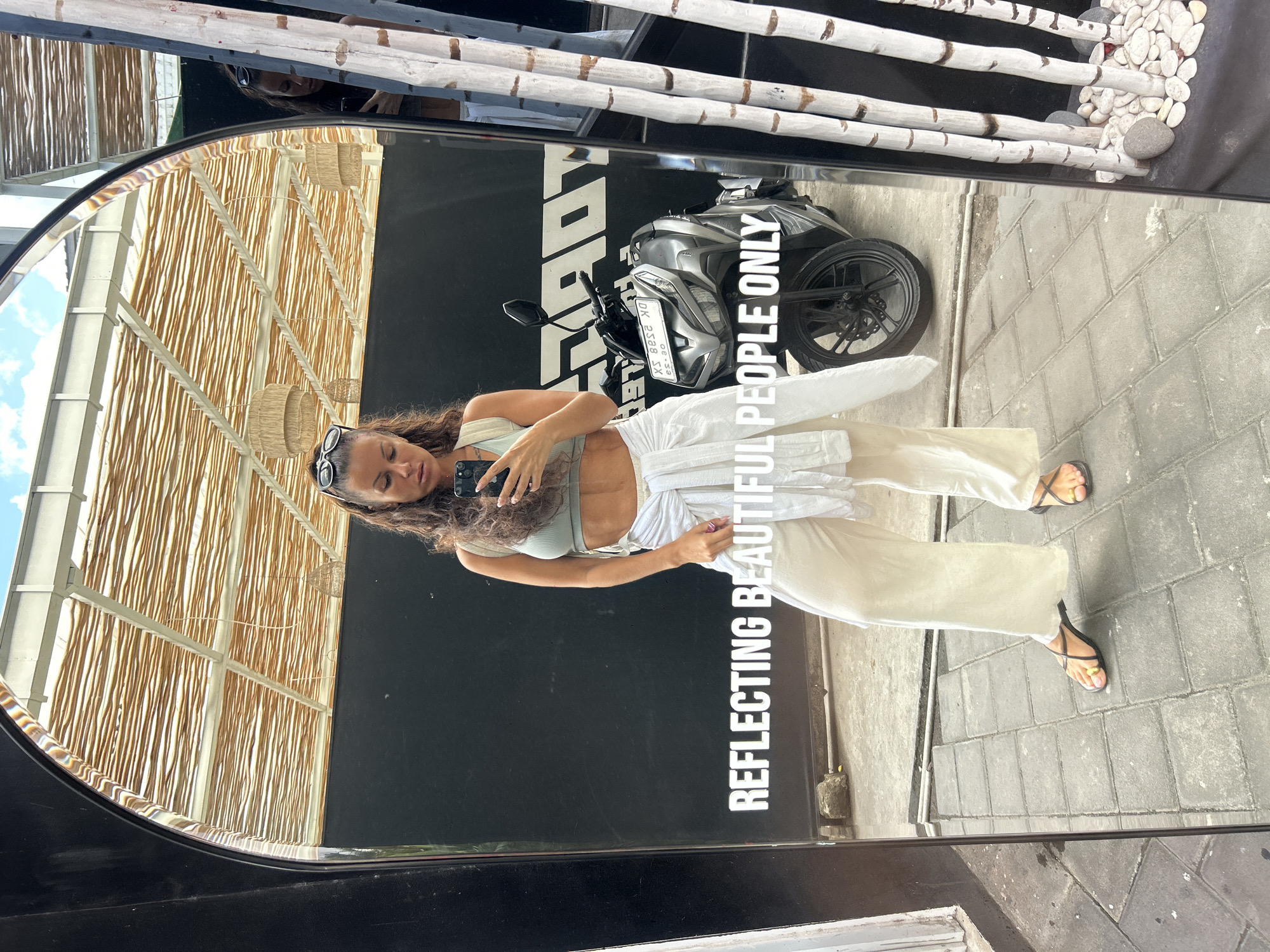 A woman is standing in front of a large mirror, taking a selfie. She is wearing a light green crop top, off-white wide-leg pants, and black sandals. A white shirt is tied around her waist. Her sunglasses are perched on her head. In the reflection of the mirror, a grey motorcycle is visible. The text "REFLECTING BEAUTIFUL PEOPLE ONLY" is displayed in white lettering on the mirror. The background appears to be an outdoor area with a black wall, a tiled floor, and a roof structure with woven material. The location is identified as North Kuta, Indonesia.