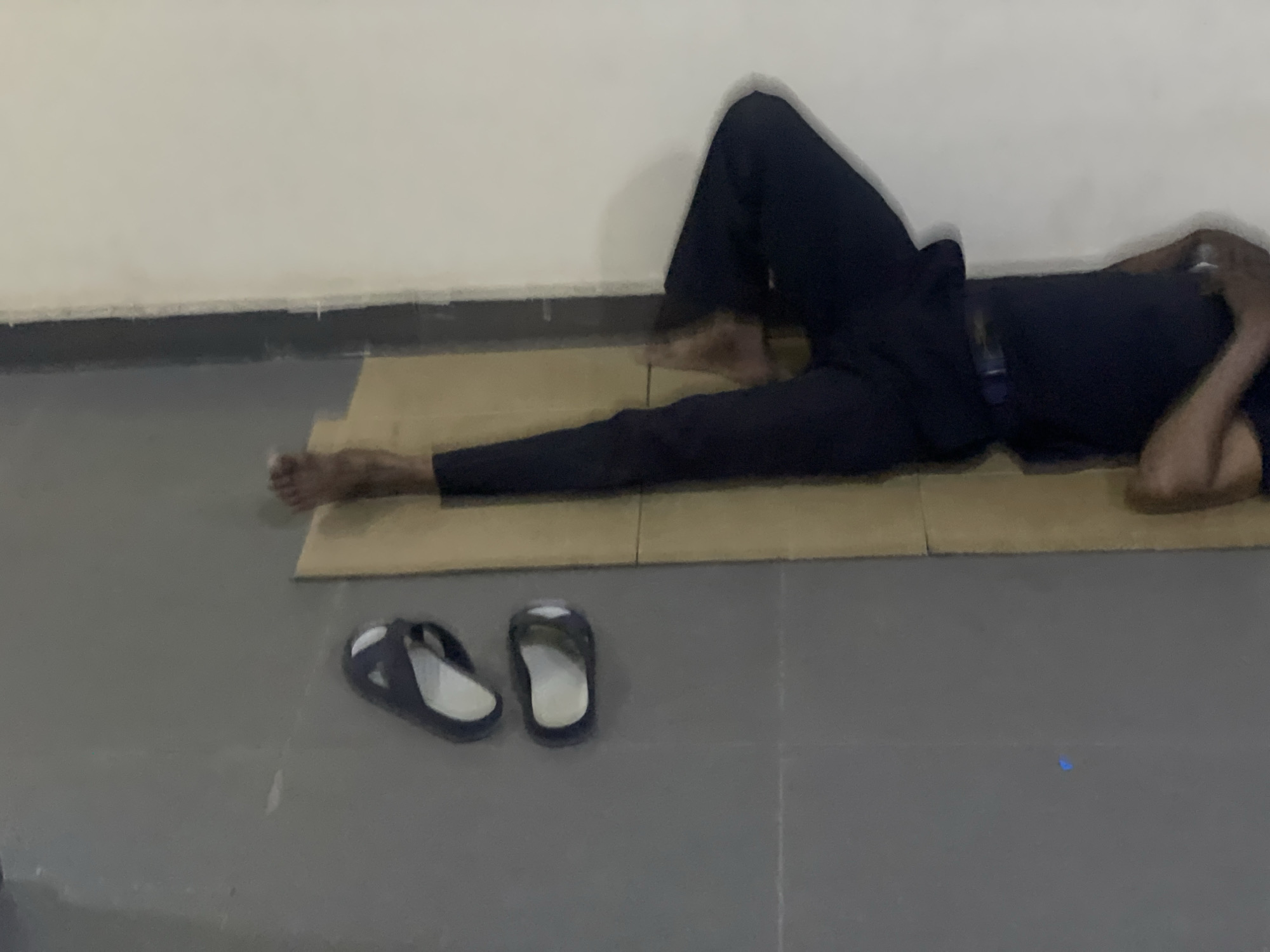 A man is lying on a folded cardboard box in a room with a white wall and gray tile floor. He is dressed in dark clothing. His sandals are near the cardboard box. The scene is likely in Abuja, Nigeria, though this is based on contextual knowledge, not visual elements.