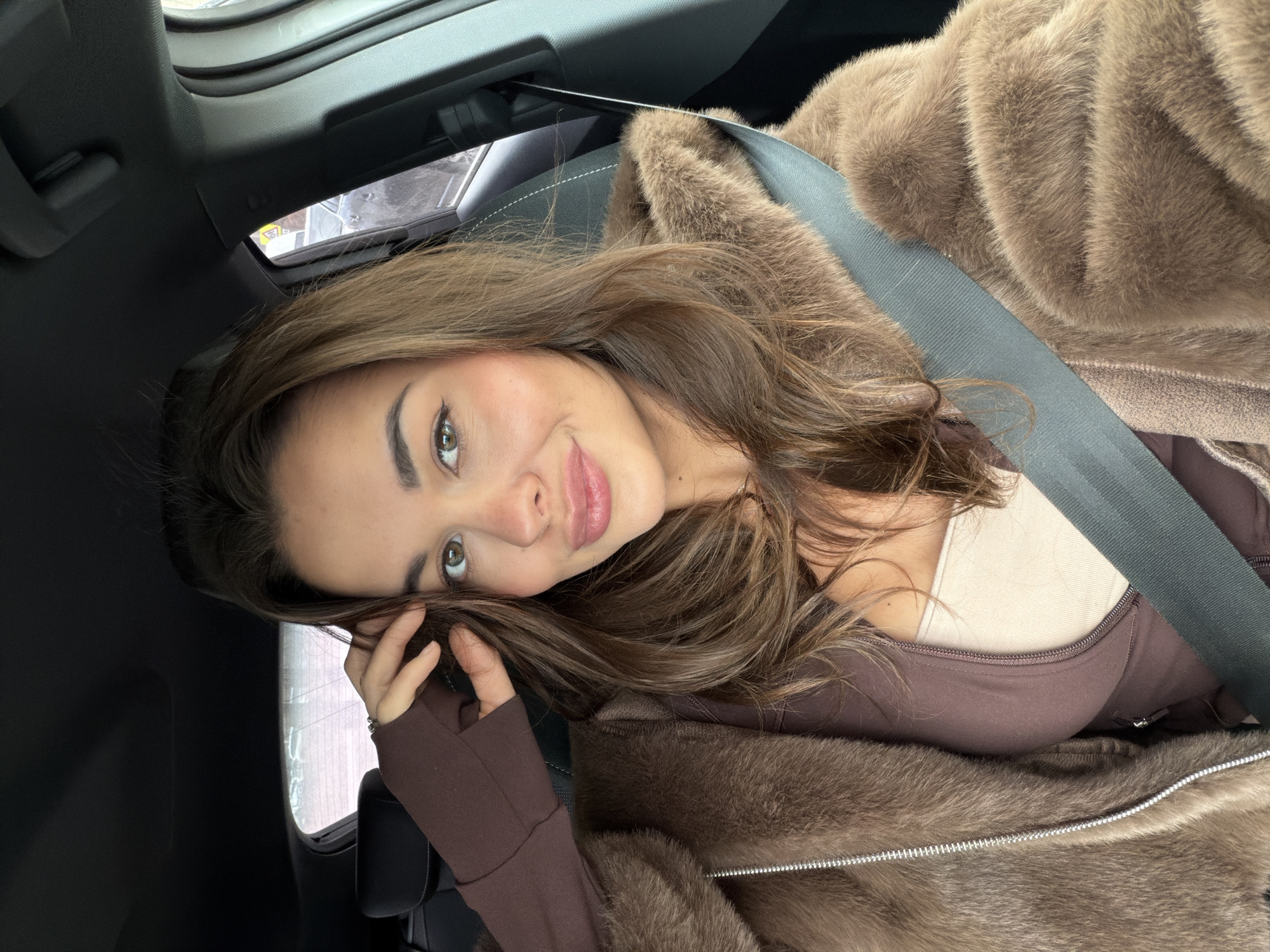 A woman is sitting in the driver's seat of a car, wearing a brown fur coat and a brown top with a cream-colored undershirt. She is wearing a seatbelt. Her left hand is near her face, touching her hair. The car's interior is visible, including the ceiling and part of the window. The exact location within Krasnoyarsk, Russia cannot be determined from the image.