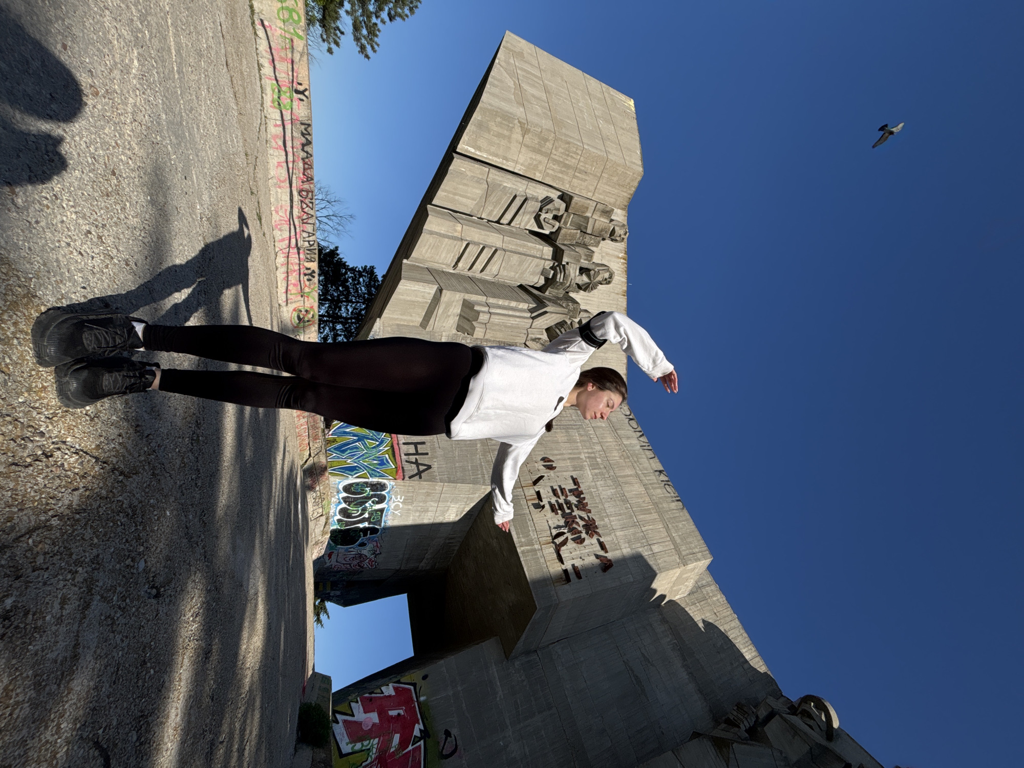An outdoor photograph, rotated 90 degrees counter-clockwise, captures a young female posing in a horizontal "flying" position against a prominent brutalist monument under a clear blue sky.
The main subject, a young woman, is dressed in black leggings, a white long-sleeve top, and black sneakers. She extends her body parallel to the ground (which appears as a vertical wall due to the rotation), with arms outstretched as if suspended mid-air. Her distinct shadow is cast on the paved surface below.
The background features a large, angular concrete monument with textured, sculptural elements, typical of Soviet-era brutalist architecture. This massive structure and surrounding concrete walls are adorned with colorful graffiti. The ground leading up to the monument is paved.
Notable details include the bright, natural daylight, indicating a clear and sunny day, likely morning or afternoon, given the strong shadows. A single bird is visible flying in the expansive blue sky. Visible text includes "МАЛАДА БЪЛГАРИЯ" (Young Bulgaria) and "HA" on a graffiti-covered wall near the subject's feet, and "CALVIN KLEIN" and a stylized skeletal figure in graffiti on the monument itself. The setting, with its distinctive architecture and graffiti, is located in Varna, Bulgaria.