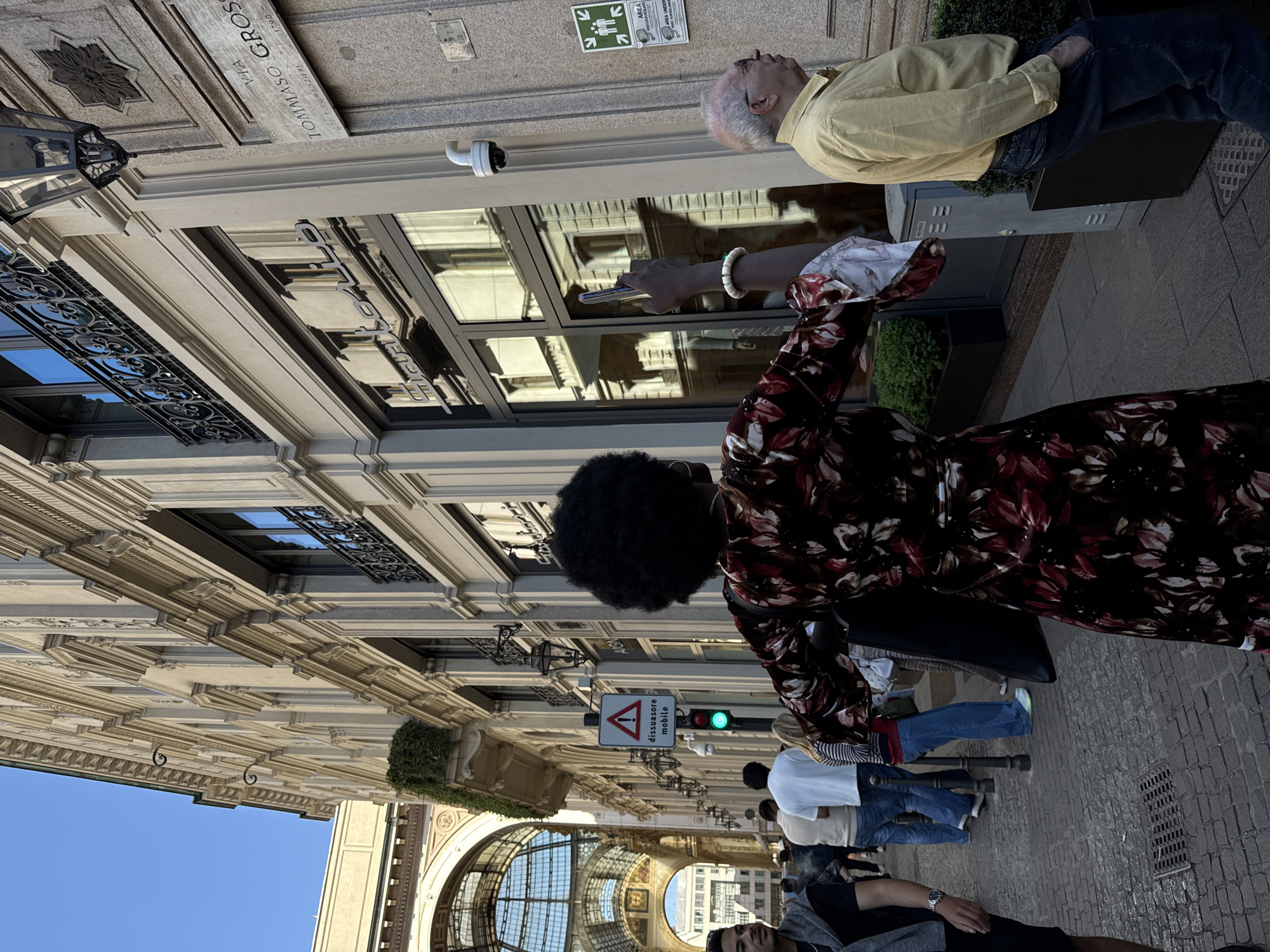 The image is taken at the Galleria Vittorio Emanuele II in Milan, Italy. The perspective is from a low angle, looking up at the architectural details of the Galleria. The scene includes a mix of pedestrians. A woman is holding a smartphone. A sign with the text "dissuasore mobile" is visible. The sky is a clear blue. The surrounding buildings are ornate and detailed.