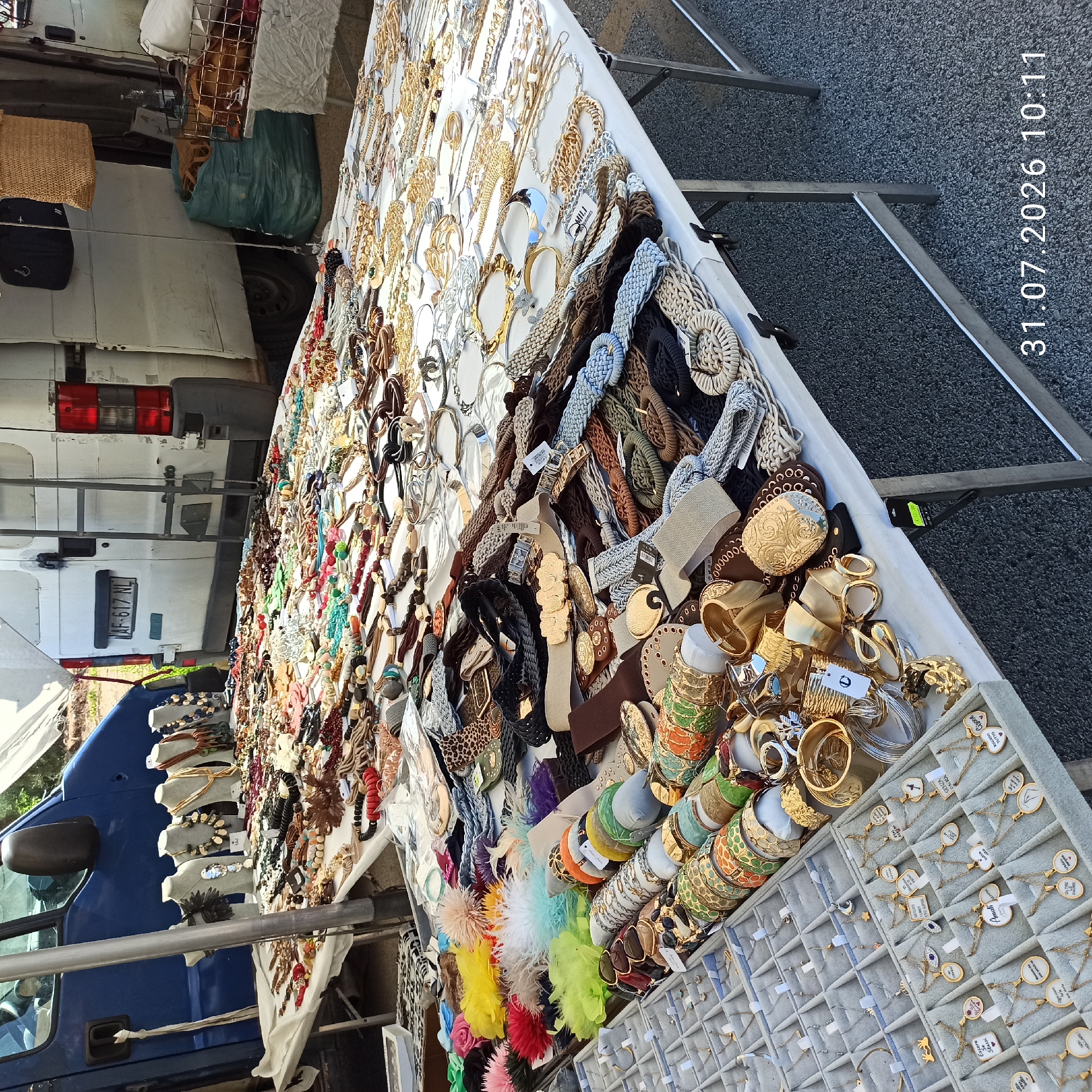 In Modugno, Italy, an outdoor market stall displays a wide assortment of jewelry. The stall is laden with necklaces, bracelets, earrings, and rings, many of which are gold or silver-toned. Some pieces feature colorful beads or stones, while others are more minimalist in design. The jewelry is arranged on white cloths and in display trays, with some items also presented on mannequins. Behind the stall, a white van is parked, with a blue vehicle partially visible to the left. The ground is covered in asphalt. The scene is illuminated by natural daylight.