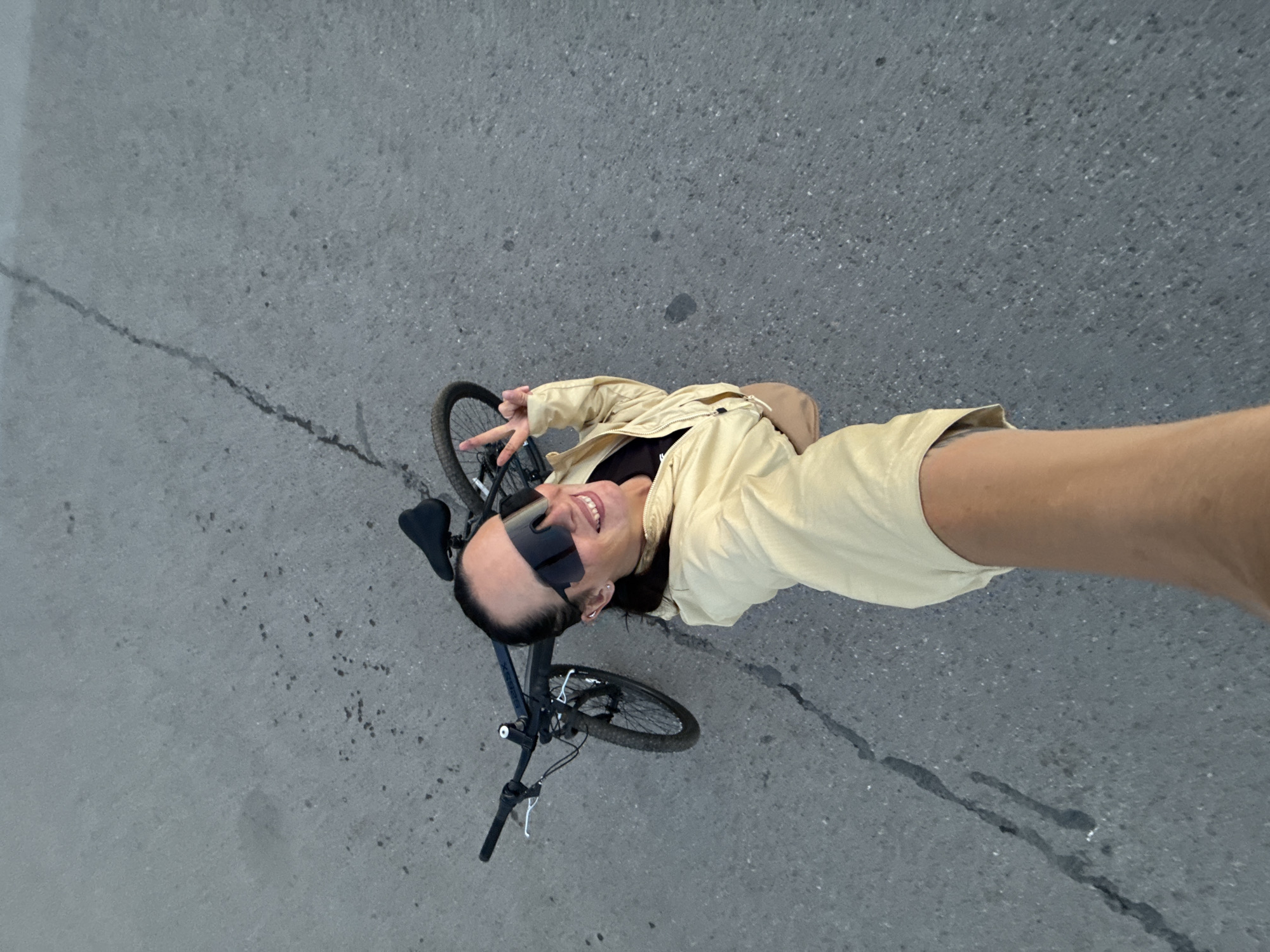 A woman, wearing a light yellow jacket, sunglasses, and black top, takes a selfie with a bicycle. She is smiling and holding up two fingers in a peace sign. The bicycle is dark colored and leaning against the ground. The scene takes place on a cracked asphalt road in Chaikovsky, Russia.