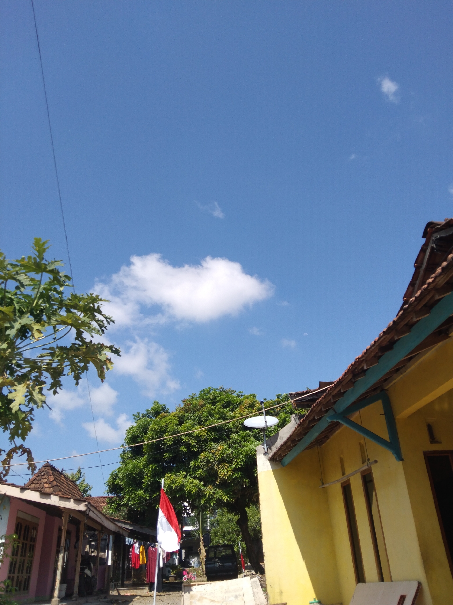 The image depicts a residential scene in the city of Kelet, Indonesia, under a clear blue sky dotted with a few white clouds. In the foreground, a tree with large, green leaves is visible on the left, its branches extending towards the sky. A thin black wire runs diagonally across the upper left portion of the frame. On the right, the corner of a yellow house with a tiled roof is prominent, featuring a light blue trim under the eaves. A window with wooden framing is visible on the yellow house. Further into the scene, to the left, a smaller pink house is partially visible, also with a tiled roof. In front of the pink house, a flag with red and white horizontal stripes, the Indonesian flag, is displayed on a pole. Clothing is hanging on a clothesline between the houses. A large green tree with dense foliage stands behind the flag and between the houses. A satellite dish is mounted on the wall of the yellow house. The ground appears to be unpaved. In the background, other houses and trees are discernible, suggesting a village or suburban setting. The lighting indicates it is daytime, likely late morning or early afternoon, given the bright sunlight casting shadows.