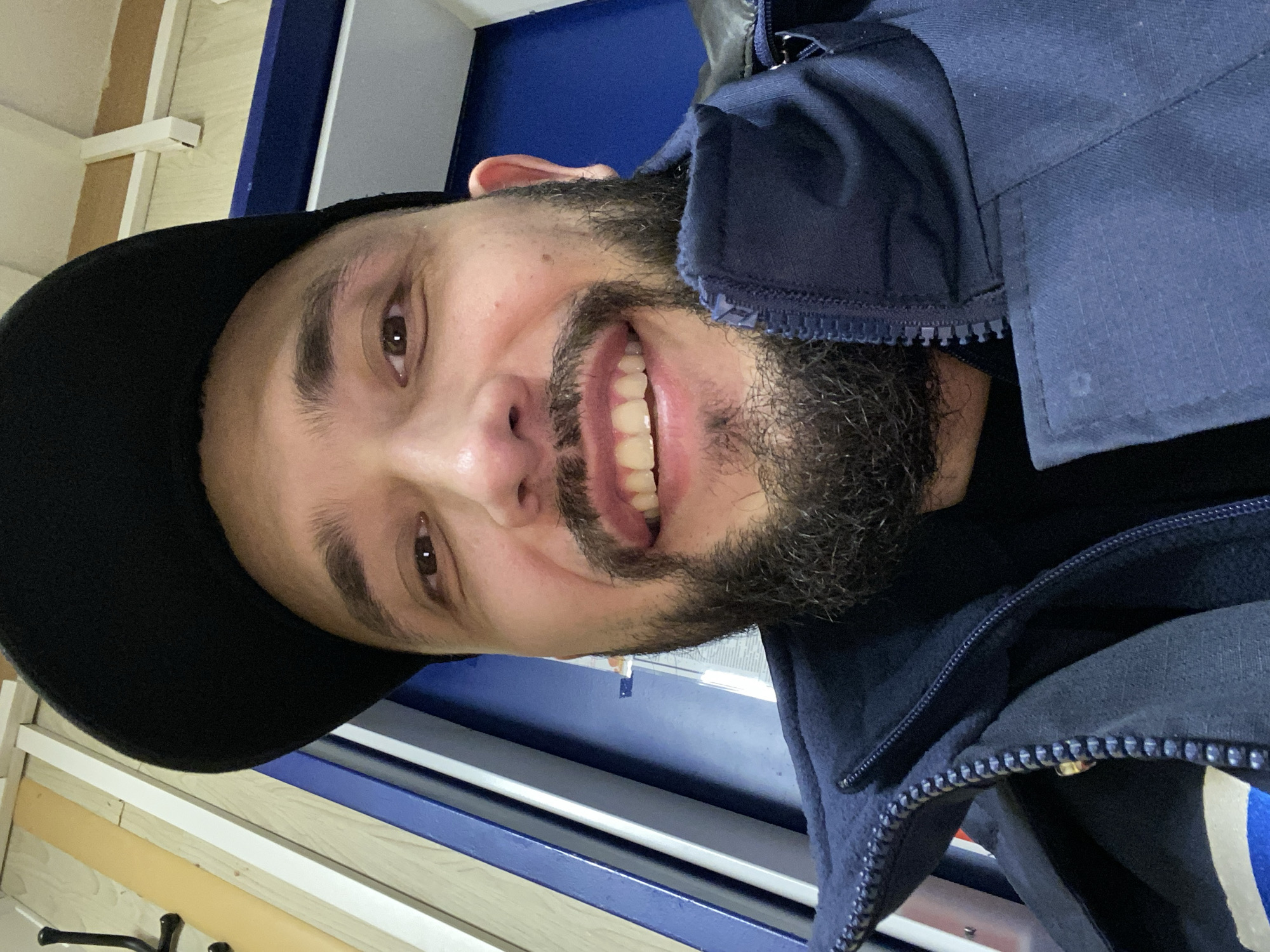 A close-up, rotated portrait shows a man smiling broadly at the camera. He has a dark, full beard and mustache, and dark brown eyes. The man is wearing a black baseball cap and a navy blue zippered jacket, which appears to have a fleece-like lining or collar. A dark garment is visible beneath the jacket.
The scene is indoors, likely under artificial lighting. The background features light-colored wooden or laminate panels and a white trim to the man's left. To his right and behind him, a prominent dark blue surface is visible, possibly a wall, a large piece of furniture, or equipment. The man's expression is joyful and friendly. There is no visible text in the image. The location is contextualized as Koskolovo, Russia.