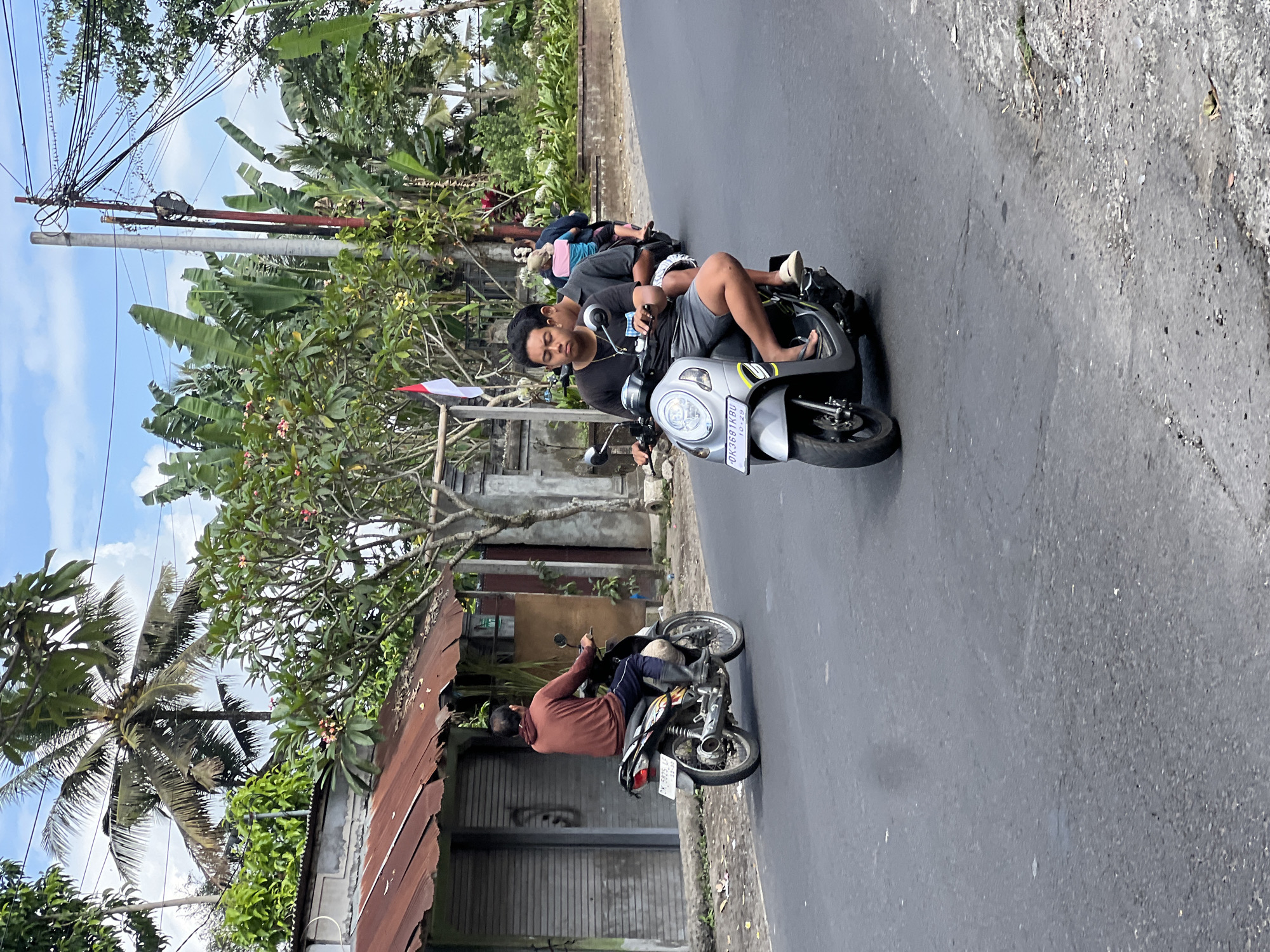 The image depicts a street scene in Payangan, Indonesia, with several individuals riding on motorcycles. In the foreground, two young men are on a silver scooter, with the driver wearing a black t-shirt and grey shorts, and the passenger in a dark grey t-shirt. Behind them, another scooter carries two individuals, one in a blue and pink shirt and the other in a darker shirt, both appearing to be male. Further down the road, a man in a reddish-brown hooded shirt and dark pants is riding a motorcycle. The environment includes a paved road with some debris, a building with a corrugated metal roof on the left, and lush green vegetation including palm trees and banana trees on the right and in the background. A utility pole with numerous electrical wires is also visible on the right. The sky is partly cloudy.