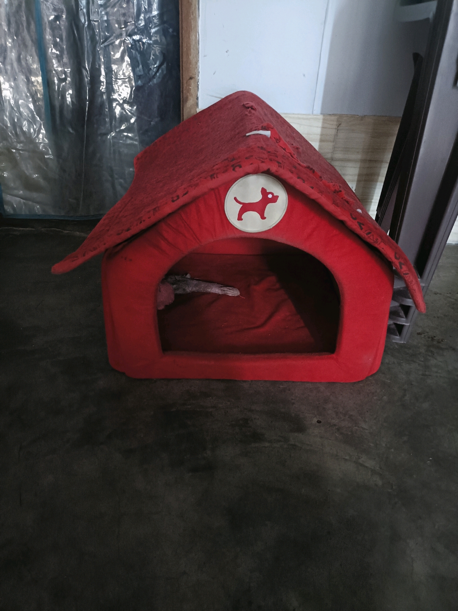 A red dog house with a white dog silhouette logo sits on a dark concrete floor. The dog house has a peaked roof and an arched entrance. Inside the dog house, a light-colored object, possibly a toy, is visible. To the left of the dog house is a large sheet of shiny, crinkled plastic, and behind it, a wooden structure. To the right of the dog house is a dark gray, multi-tiered object. The overall scene appears to be in an indoor, possibly unfinished or storage area. There are no people or animals present. The image is a full-frame shot of the dog house and its immediate surroundings.