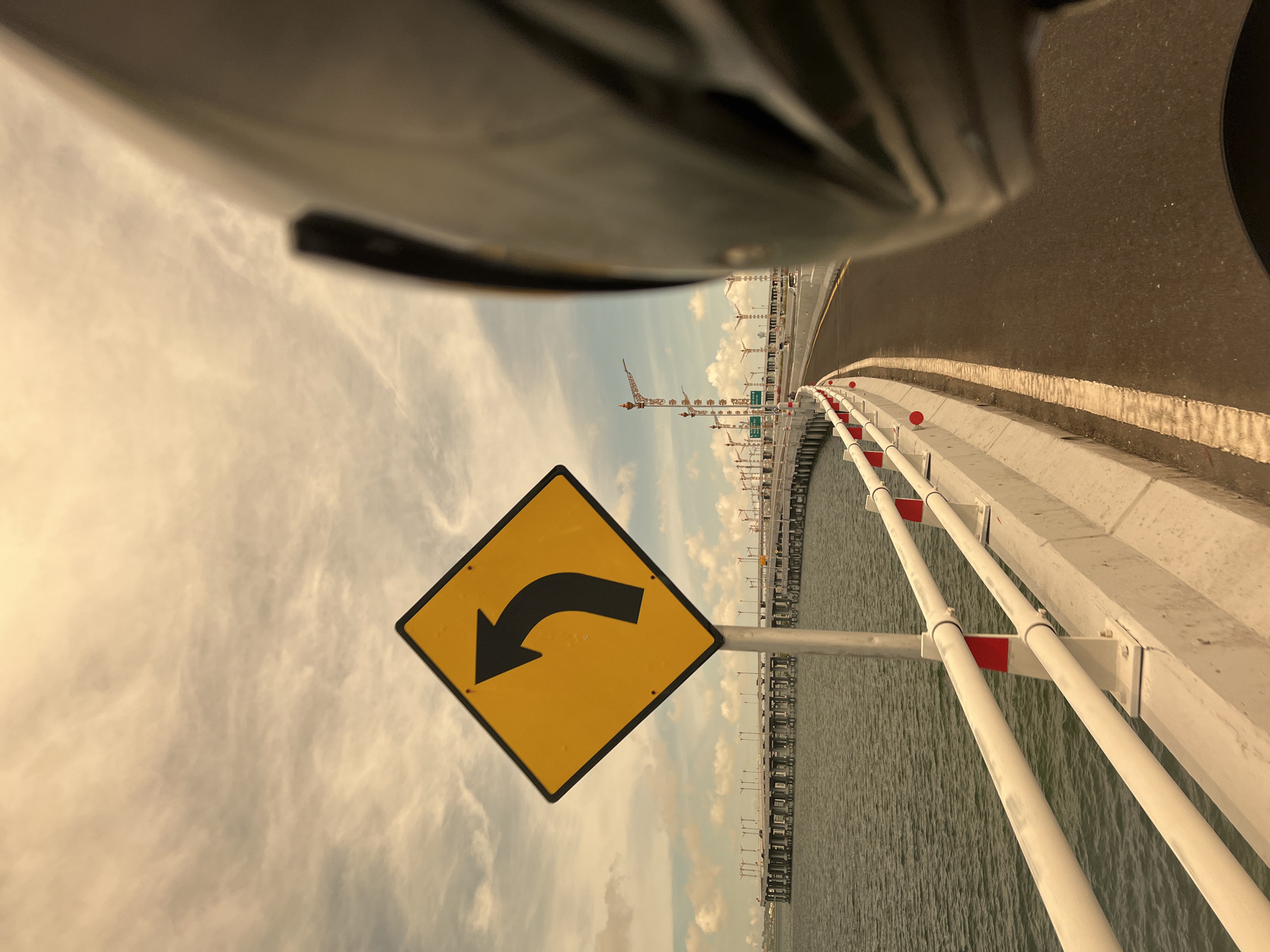 The image depicts a yellow and black road sign warning of a left turn on a bridge. The sign is situated on the left side of a road that stretches across water. The bridge has a white concrete barrier with metal piping along the side. Cranes are visible in the distance along the shoreline. The sky is overcast with clouds. The location appears to be the Mandara Toll Road in Kuta, Indonesia, as the scenery matches that location, including the bridge architecture and distant industrial activity. The image appears to be taken from a vehicle or motorcycle, with part of the vehicle visible in the upper portion of the frame.