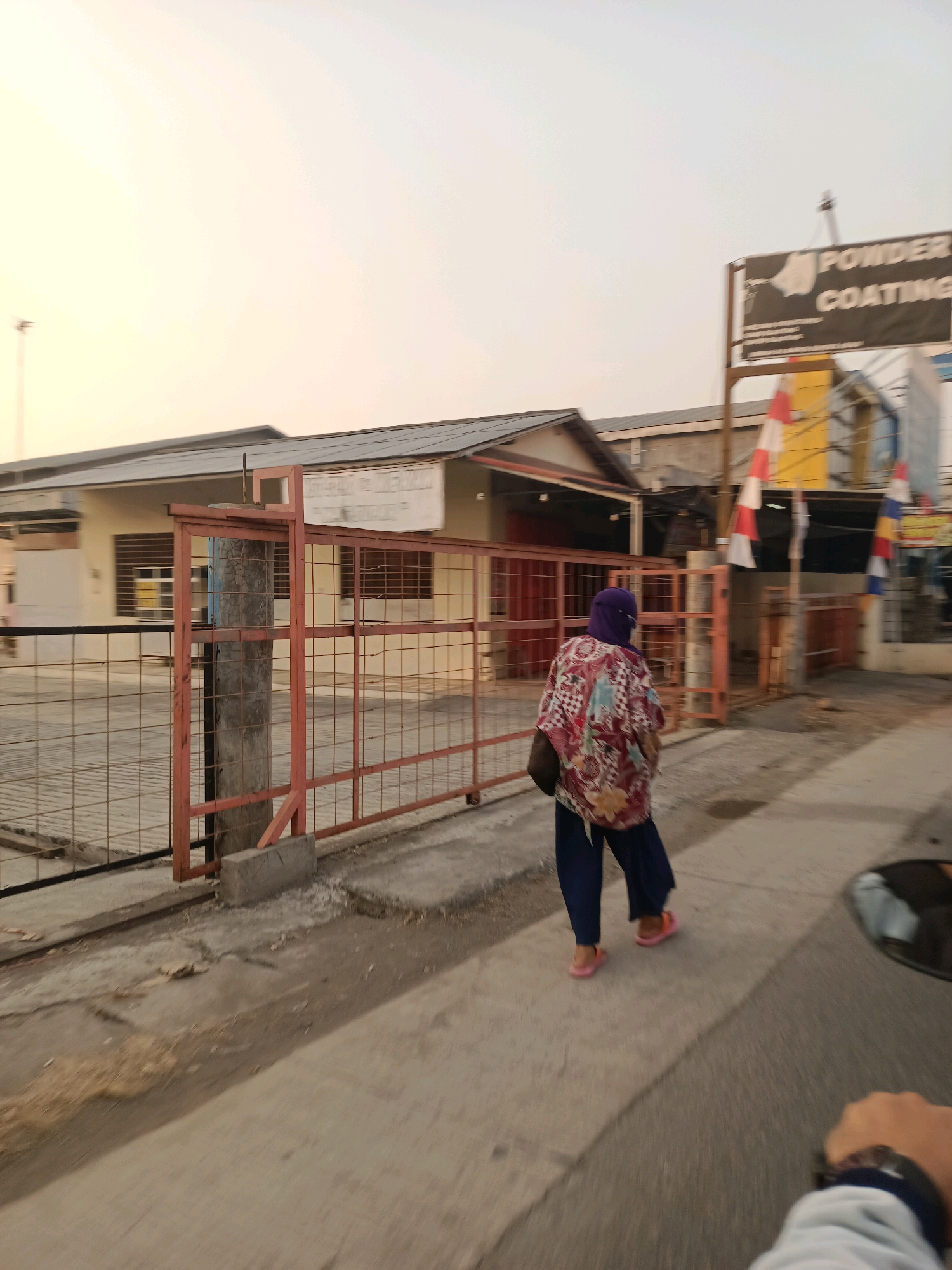In Karawang, Indonesia, a person walks along a paved sidewalk in front of a fenced industrial property. The individual, wearing a headscarf, a patterned shirt, and dark pants, walks away from the camera, towards the right side of the frame. A sign visible above the fence reads "POWDER COATING". The buildings in the background appear to be industrial or commercial in nature. The sky is bright and overcast, suggesting either early morning or late afternoon lighting conditions. The foreground shows a portion of a road or pathway, with some debris scattered on the ground.