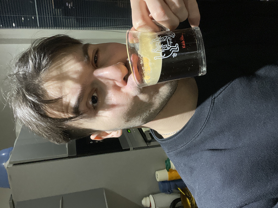 A person drinks from a glass mug with a Keith Haring design. The mug contains a dark liquid with a light-colored foam on top. The person has short brown hair and fair skin. A silver necklace is visible around the neck. Behind the person is a countertop with various objects, including tubes and bottles, and an appliance, likely a microwave, is behind the person's head. The background has window blinds. The image appears to be taken indoors in artificial lighting.