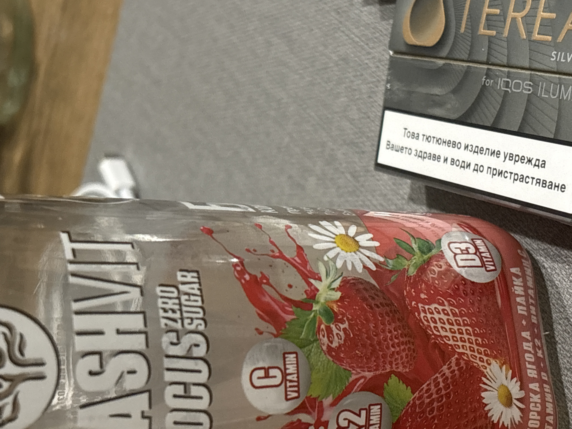 The image shows a close-up of two items: a can of ASHVIT focus drink (zero sugar, strawberry flavor) and a pack of TEREA for IQOS ILUMA (tobacco product). The ASHVIT can features images of strawberries and daisies, highlighting the presence of vitamins C, D3, and K2. The TEREA pack has a health warning in Bulgarian: "Това тютюнево изделие уврежда Вашето здраве и води до пристрастяване" ("This tobacco product harms your health and leads to addiction"). The location is likely Sofia, Bulgaria, indicated by the Bulgarian language on the TEREA pack.