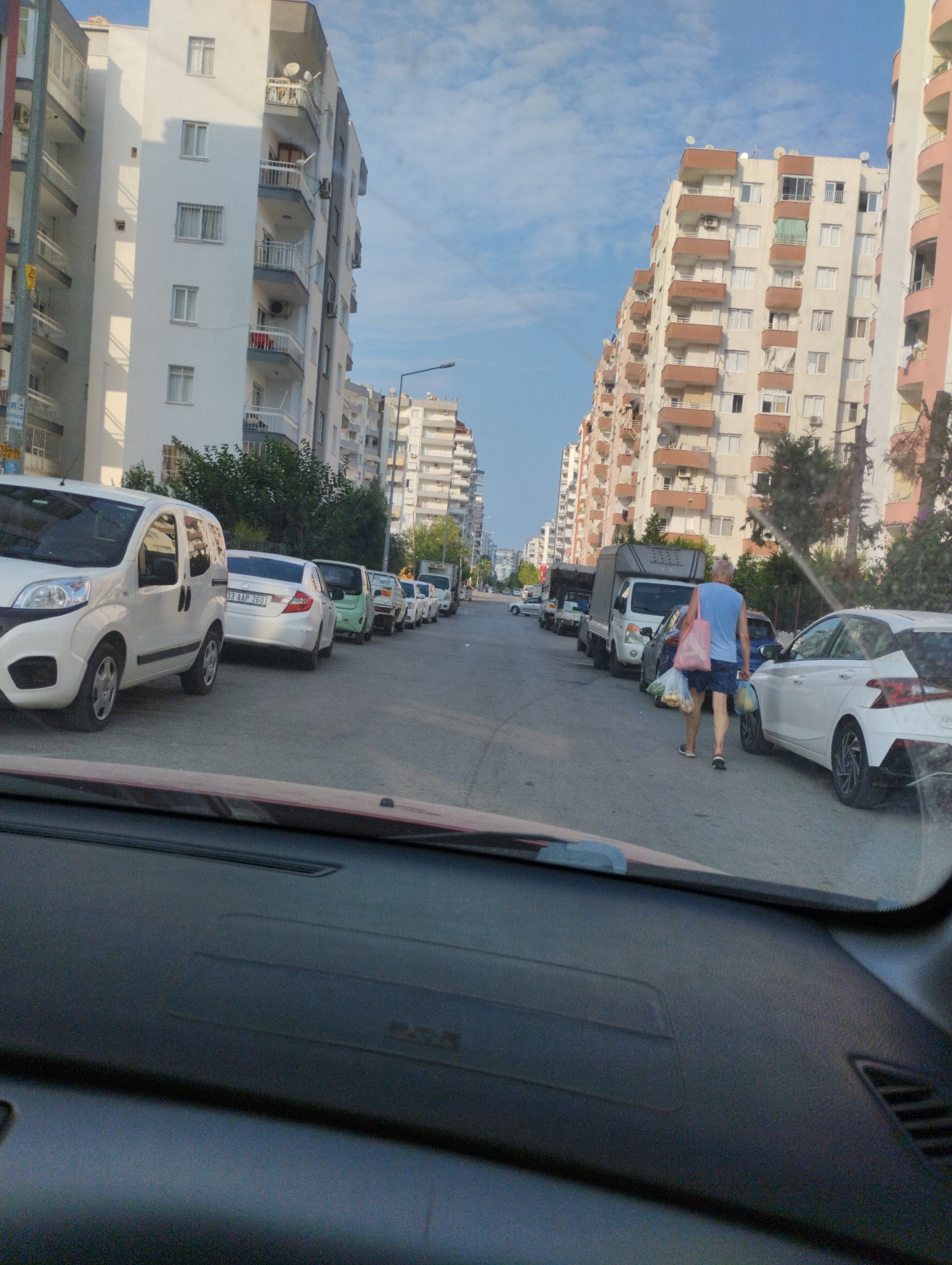 The image shows a street scene in Mezitli, Turkey. On the left side of the street are several parked white cars, including a Fiat Fiorino. Further down the street, there are more parked cars and buildings. On the right side of the street, a man is walking away from the camera, carrying two shopping bags, one pink and one white, filled with groceries. He is wearing a blue t-shirt and dark shorts. A white car is parked next to him. The street is lined with multi-story apartment buildings. The sky is blue with some clouds. The image appears to be taken from inside a car.