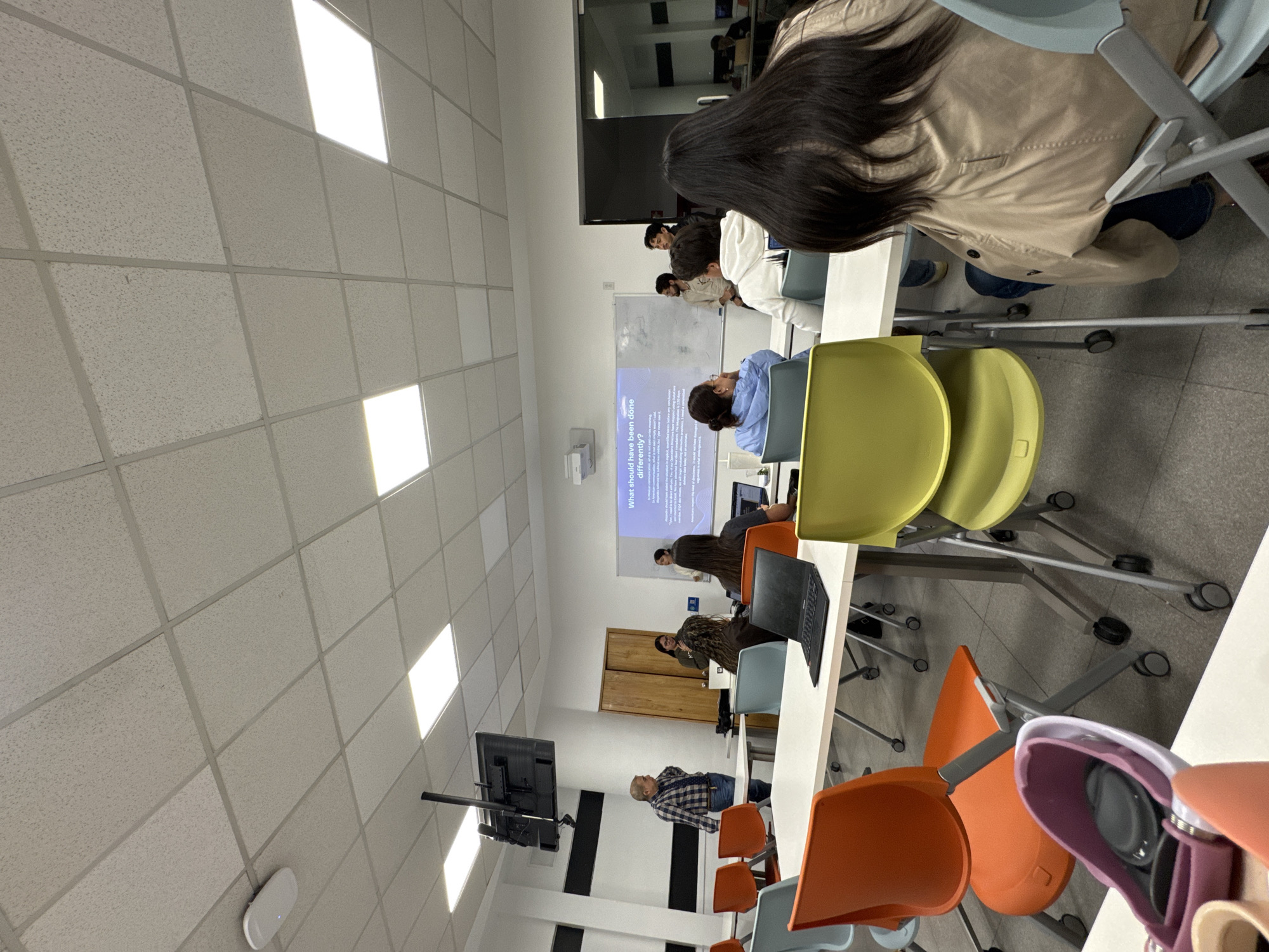 The image shows a classroom in what appears to be a modern building. The room has a suspended ceiling with integrated square light panels. The walls are painted in a light color. Students are seated at long white tables, facing a projection screen displaying text. Some students have laptops open. There are colorful chairs, including orange, yellow, and light blue ones. A flat-screen display is mounted on the wall at the front of the classroom. Based on visual features and likely campus context, the location is Puebla, Mexico.