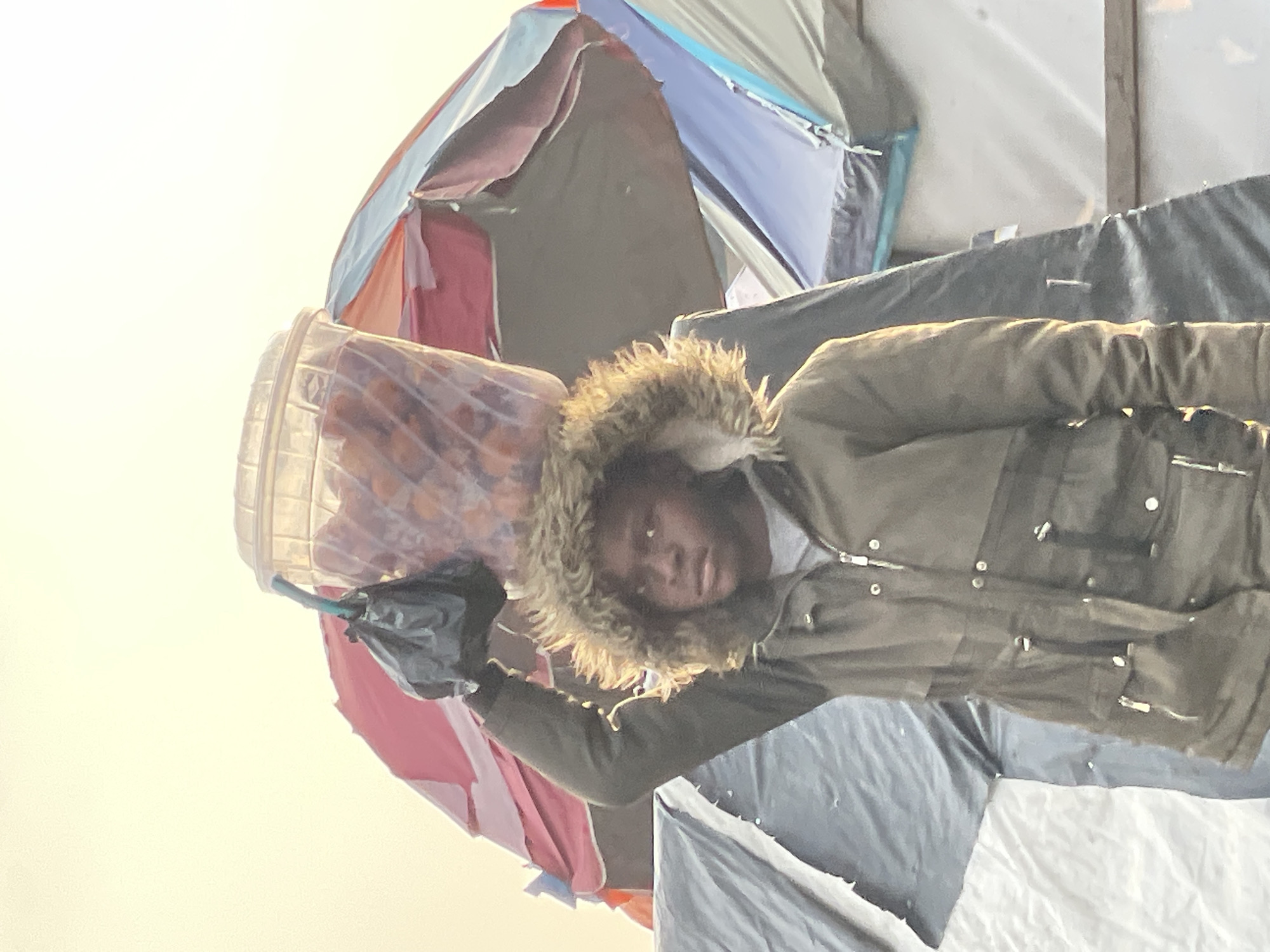 A young person, appearing to be a male, stands outdoors and carries a large, transparent plastic container filled with small, round, orange objects on their head. The container is covered with a clear plastic lid, and a black plastic bag is tied to the handle of the container, dangling down. The person is wearing a dark green hooded jacket with a fur-lined hood. Their right arm is raised, supporting the container. In the background, there are several tents and what appear to be umbrellas, suggesting an outdoor market or encampment. The lighting suggests it is either early morning or late afternoon, with a bright, overcast sky. The scene is likely in Ilado, Nigeria, an area known for its informal markets and densely populated settlements.