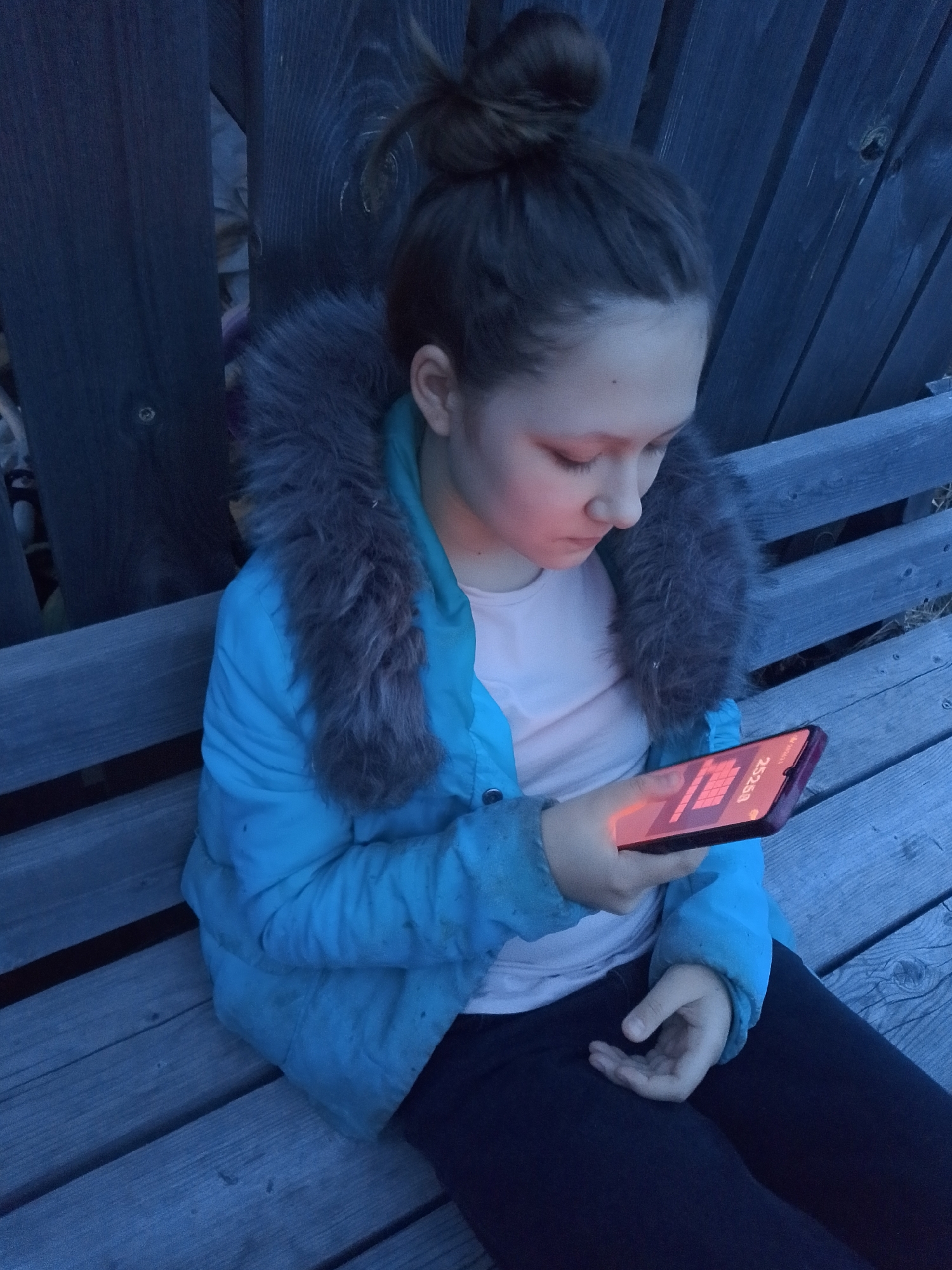 A young person with their hair in a bun sits on a wooden bench outdoors, their face illuminated by a mobile phone they are holding. The person is wearing a blue jacket with a fur-trimmed hood and dark pants. The background includes a wooden fence. The image was likely taken in the early evening or at night, based on the lighting. Based on the filename, the location is Прикамский, Russia.