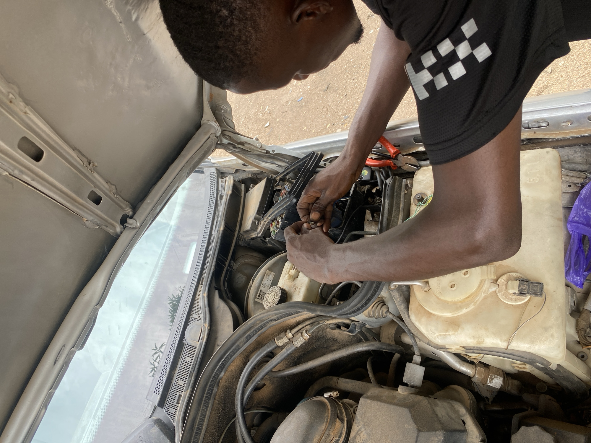In Jalingo, Nigeria, a person is seen working on a car engine under an open hood. The individual, whose head and shoulders are visible from the side, is focused on connecting wires within the engine compartment. Red-handled pliers are placed on the edge of the engine bay, suggesting tools are being used for the task. The engine is dusty, indicating it is either old or has been exposed to the elements. The surrounding environment appears to be an outdoor setting, with a sandy or dirt ground visible. The car itself is silver and appears to be a sedan.