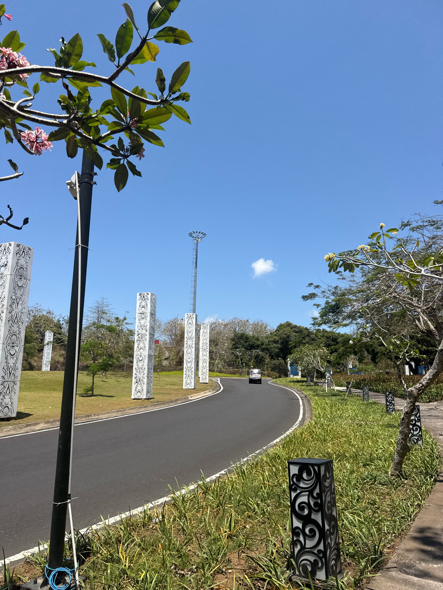 The image depicts a road curving through a landscaped area under a clear blue sky in Kuta Selatan, Indonesia. Along the left side of the road, several tall, white, rectangular pillars with intricate black patterns are erected on a grassy lawn. A black lamppost with visible wiring is positioned in the foreground on the left. Further down the road, a silver car is visible. The right side of the road features a sidewalk lined with lush green grass and decorative black lanterns. Trees with green foliage and some flowering branches are scattered throughout the scene. In the distance, a tall, metallic structure, possibly a communication tower, can be seen against the sky, which also features a single, small white cloud. The overall environment suggests an urban or semi-urban setting with a focus on decorative landscaping and infrastructure.