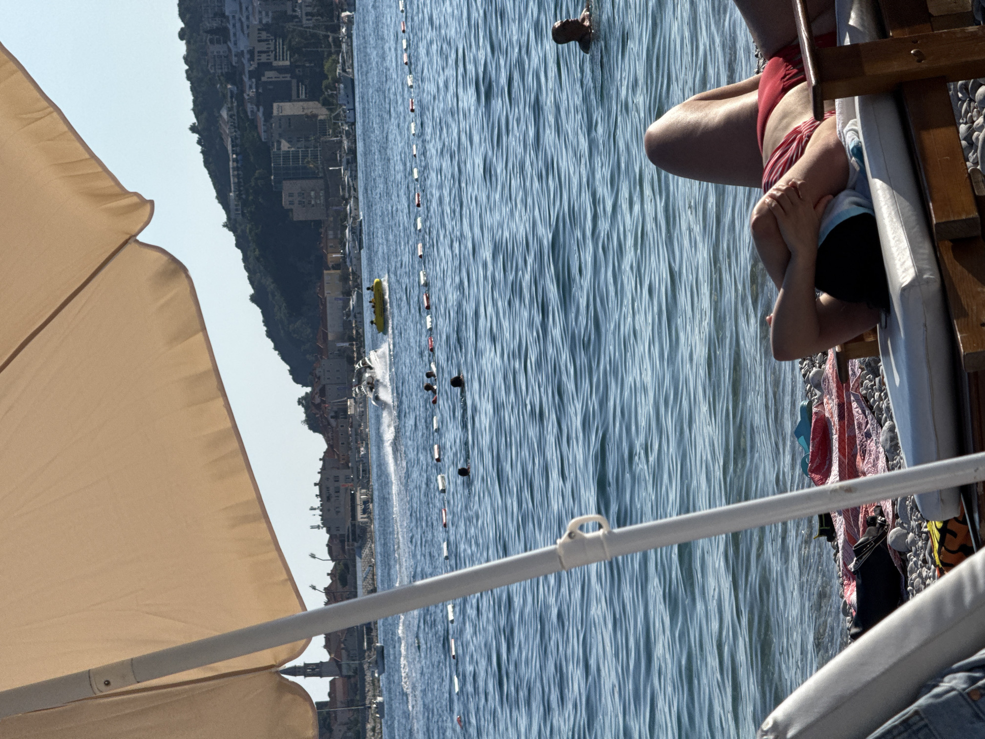 In Budva, Montenegro, on a sunny day, a person is relaxing on a lounge chair on a pebble beach, partially obscured by a large beige umbrella. The person is wearing a red and white striped bikini and has their arms behind their head. The sea is visible in the background, with several people swimming and a yellow tube being towed by a boat. A town with buildings and a church tower is visible on the coastline in the distance, situated on a hill.