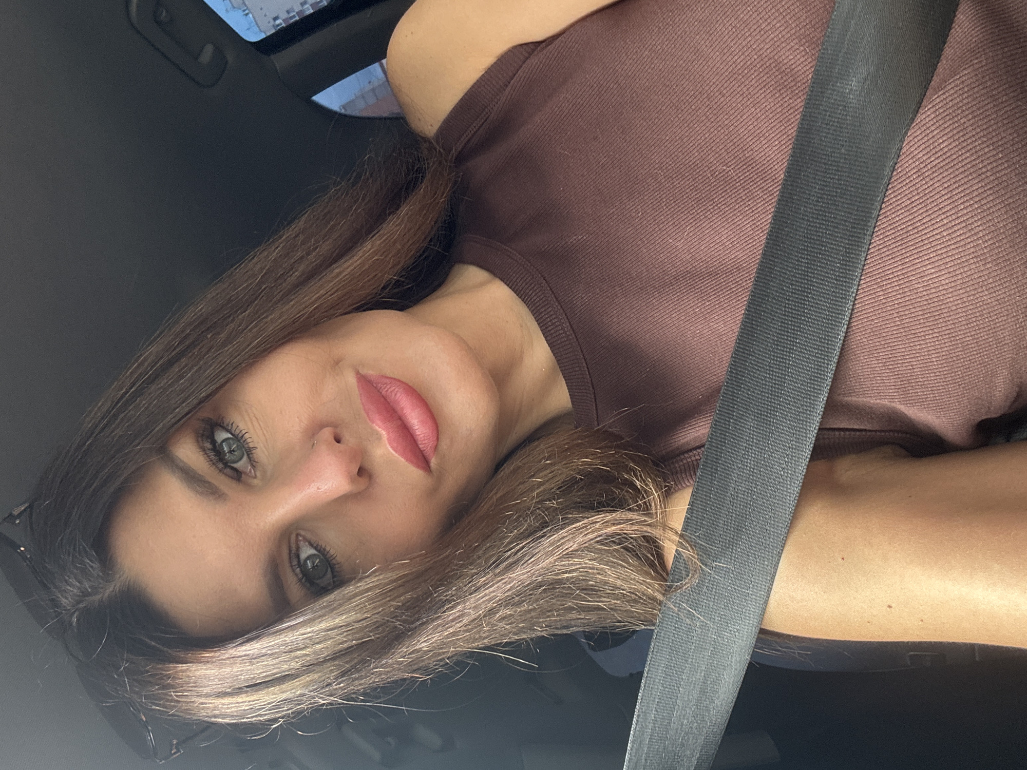 In this medium shot, a woman with long brown hair and green eyes is in the driver's seat of a car. She is wearing a brown tank top and a seatbelt. Her head is tilted slightly to the left, and she is looking at the camera with a soft smile. The background is blurry, but it appears to be a city street with buildings visible. The image was taken outdoors on a sunny day. There are no indications that this image was taken in Chaikovsky, Russia.