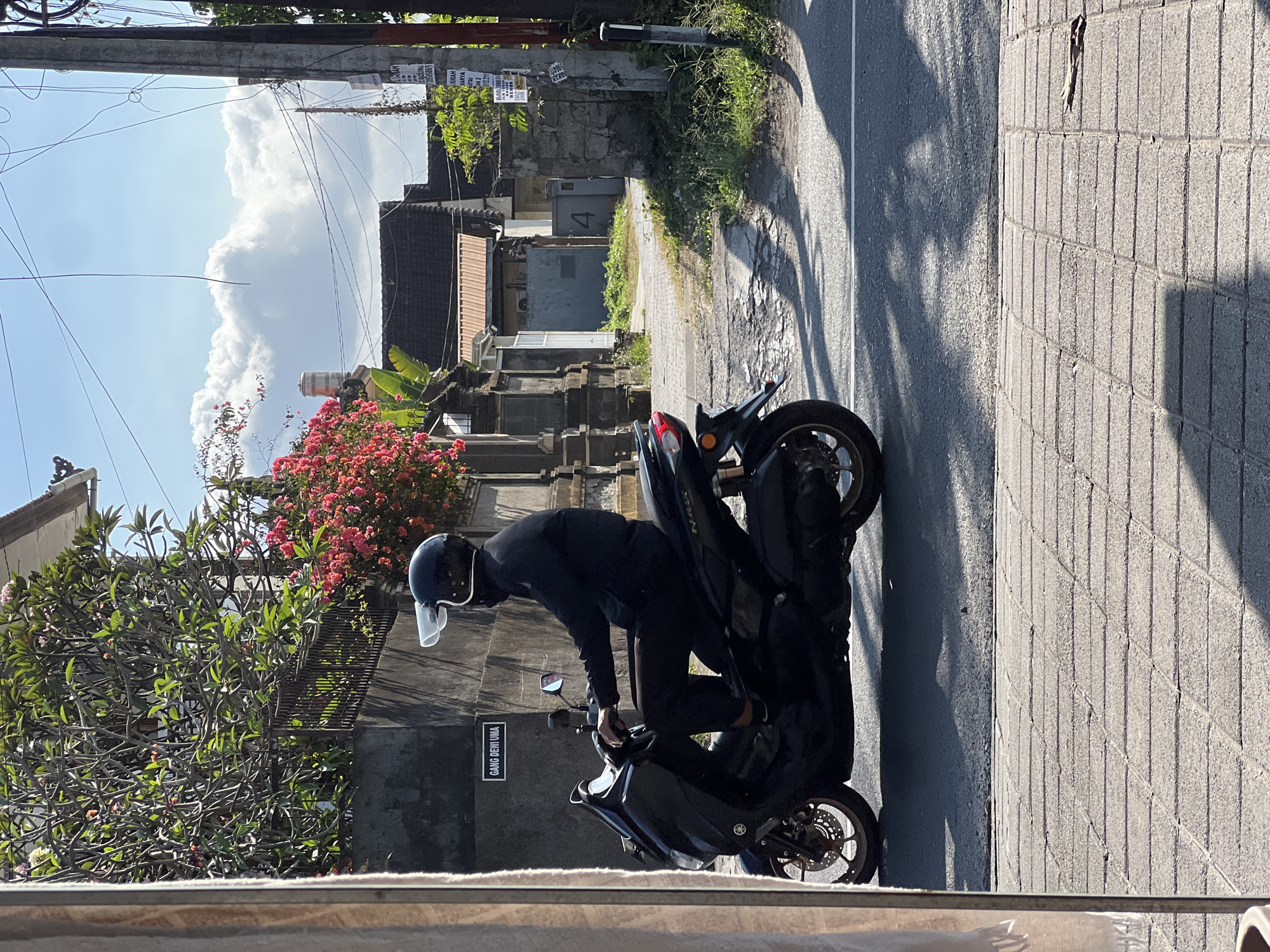 A person wearing a black helmet and all black clothing is riding a black motorcycle on a paved road in Denpasar, Indonesia. The person is seen from the side, and their face is obscured by the helmet. The motorcycle appears to be a scooter-style model. The background shows a street with buildings and trees. There is a sign on a wall that reads "GANG DEWI UMA." The sky is bright blue with some clouds. The image has a natural lighting condition, indicating it was taken during the daytime.