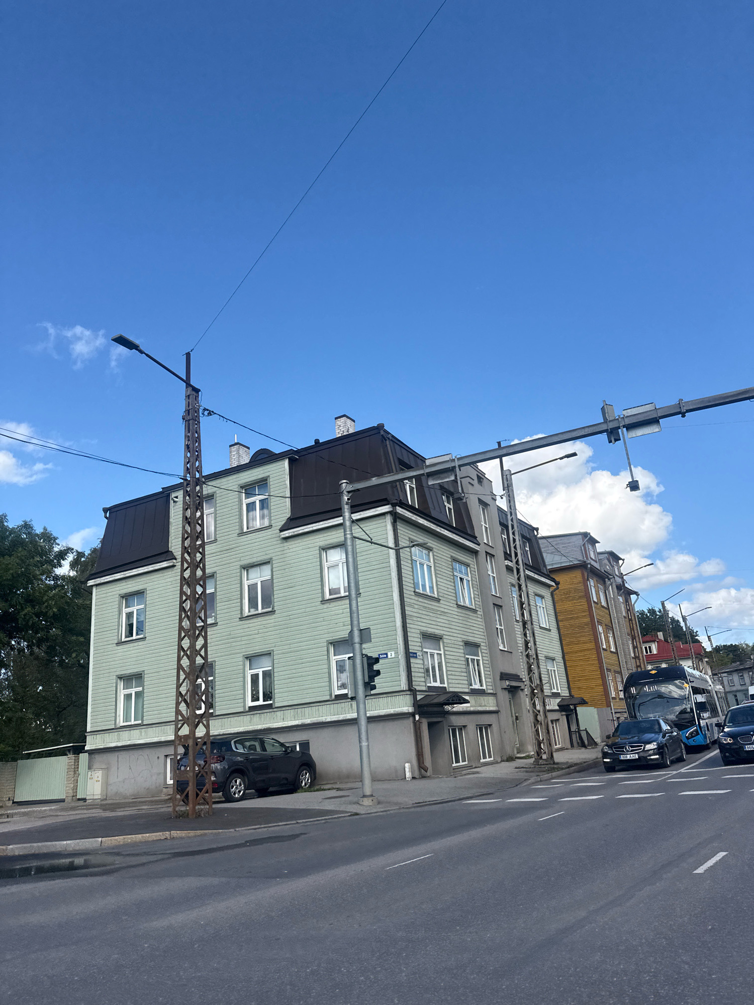 The image shows a street scene in Tallinn, Estonia, under a bright blue sky with scattered white clouds. Several multi-story buildings with pitched roofs line the street, painted in various colors including light green, grey, and yellow. A dark-colored SUV is parked on the side of the road in front of one of the buildings. Traffic signals and street lights are visible, along with power lines and utility poles. Several vehicles, including a dark sedan, a bus, and another dark sedan, are on the road. The street surface is asphalt with white lane markings. Trees are visible on the left side of the image. The overall impression is a typical urban street on a clear day.