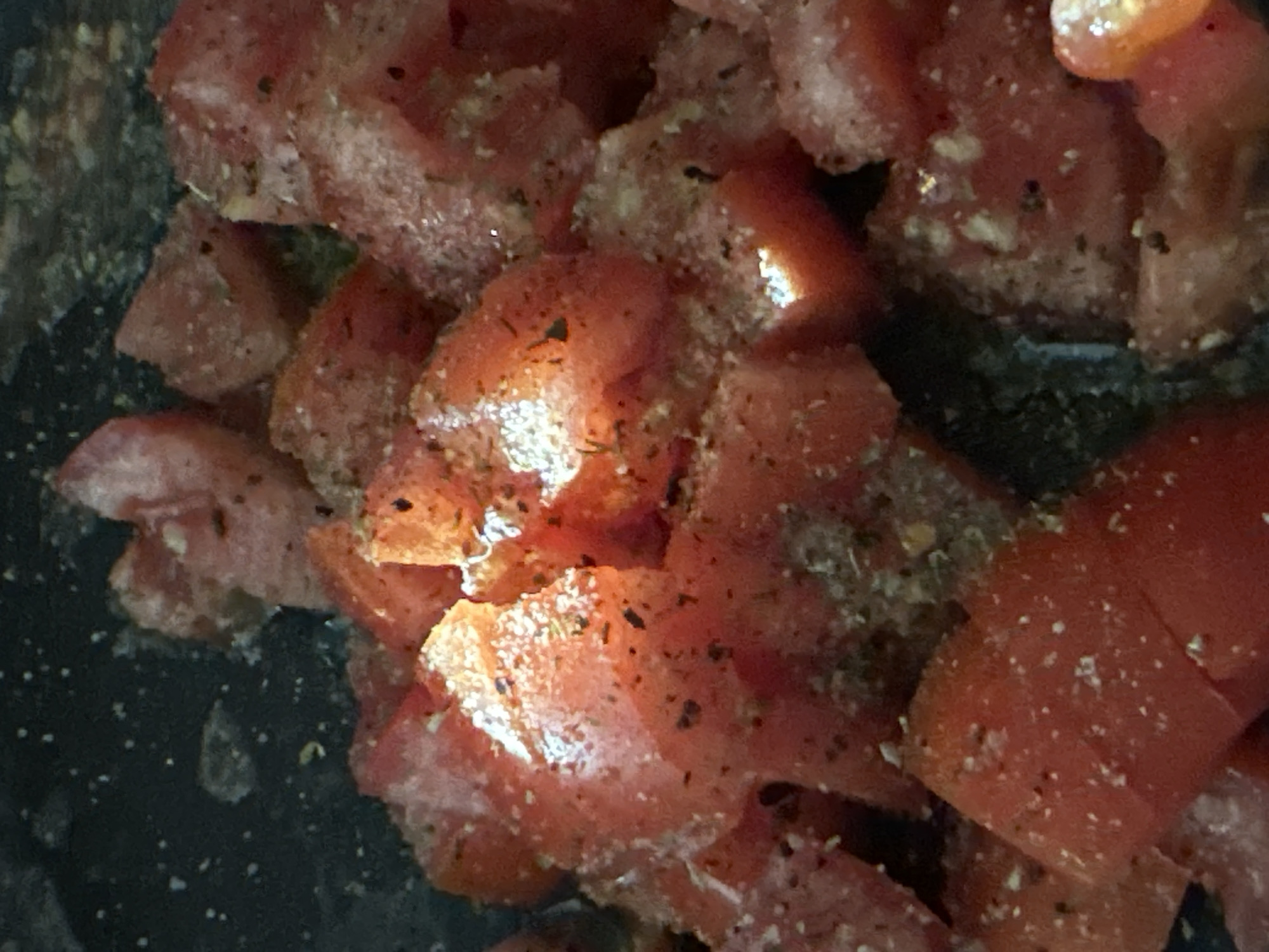 A close-up, overhead shot shows diced tomatoes, likely cooked, in a dark pot. The tomatoes are cut into irregular, roughly one-inch pieces and are coated with seasonings, including what appears to be salt and pepper. Some pieces are glistening, suggesting they are moist or have been recently cooked. The background is dark and out of focus, providing no environmental context. There are no people or animals visible in the image. The lighting highlights the texture and moisture of the tomatoes. This image does not depict any identifiable location within Krefeld, Germany.