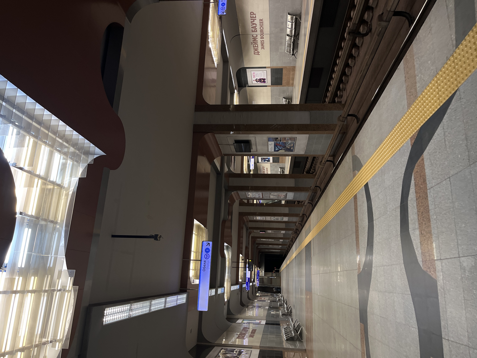 Here is a description of the image:
The image shows an underground train station platform. On the left side is a wall with arched architectural features and rows of lights. Directional signs are suspended from the ceiling, pointing towards "Обеля" and another direction. On the opposite side, the train tracks are visible. A yellow textured safety strip runs along the platform edge. The name "ДЖЕЙМС БАУЧЕР JAMES BOURCHIER" is written on the wall above some benches. There is a series of framed posters along the walls of the platform. The scene appears empty of people. The location is the "James Bourchier" station on Line 2 of the Sofia Metro.