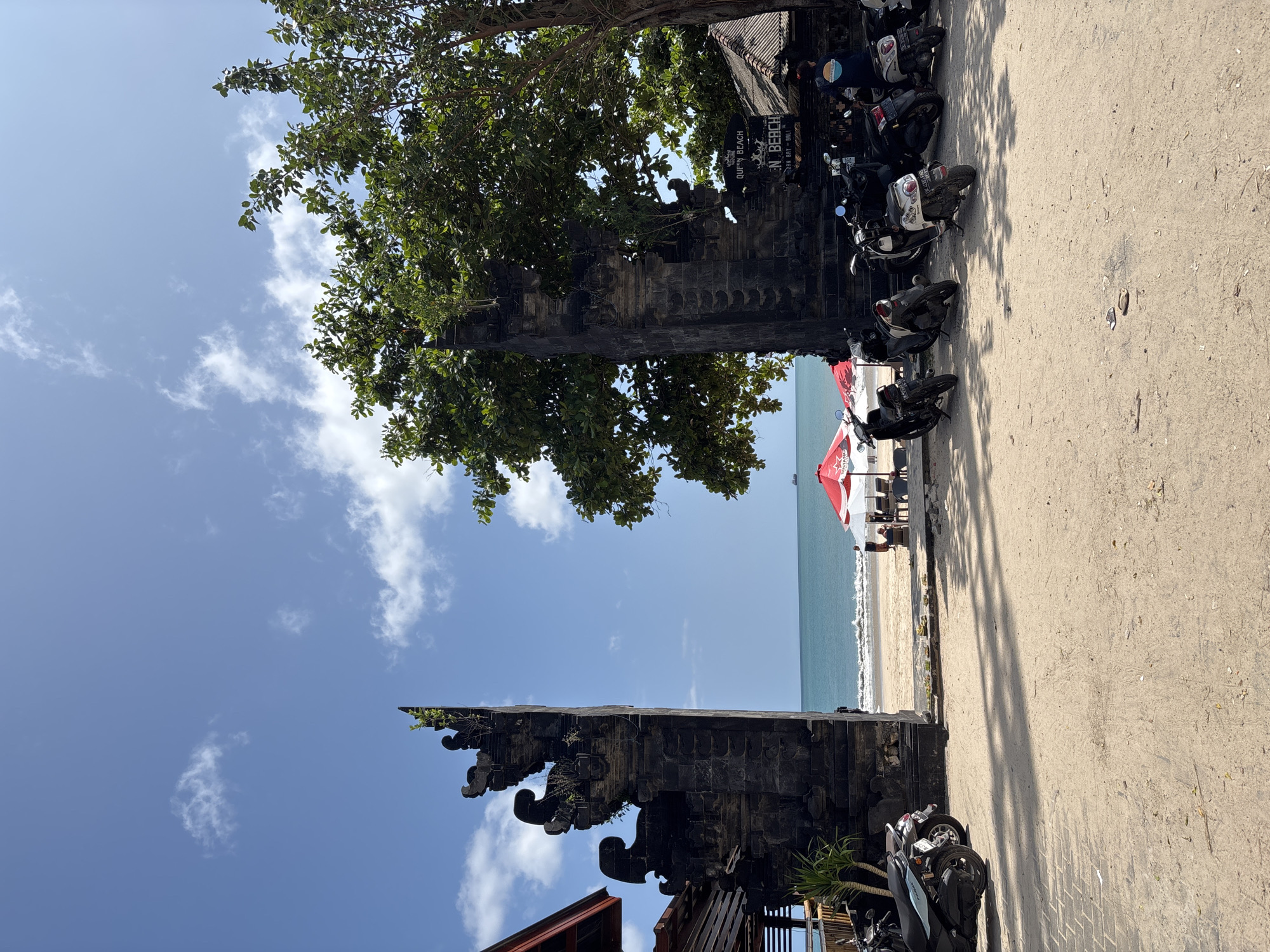The image shows a view towards the beach in Kuta Selatan, Indonesia, taken from a street-level perspective. A traditional Balinese structure, seemingly a temple gate or entrance, stands at the left of the frame, partially obscured by a tree with dense green foliage. A collection of motorbikes is parked densely along the right side of the gate. To the right of the structure, the sand of the beach is visible, with a red and white umbrella and a few people under it in the distance. The blue ocean stretches to the horizon, meeting a clear sky with scattered clouds. The overall lighting suggests it is daytime.