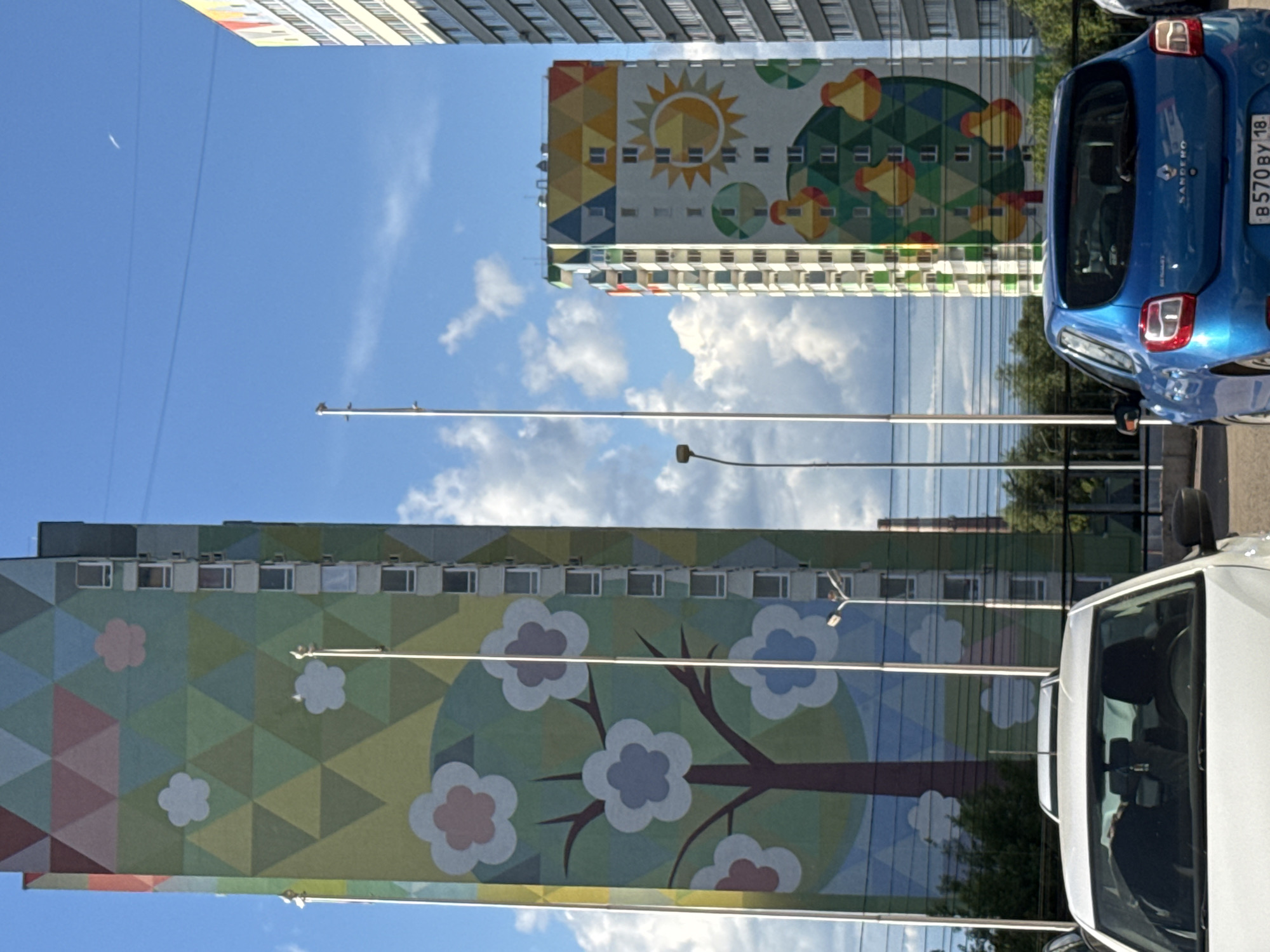 The image depicts a view looking upwards at two buildings with vibrant, colorful murals in Izhevsk, Russia. The building on the left features a large mural of a tree with flowers, rendered in shades of green, blue, and pink. The building on the right has a mural of a sun with geometric patterns. Streetlights are visible against the backdrop of a blue sky with scattered clouds. Two cars are parked on the right side of the frame: a blue Renault Sandero with the license plate "B570 BY 18" and a white car. The image is taken from a low angle, emphasizing the height of the buildings and the details of the murals.