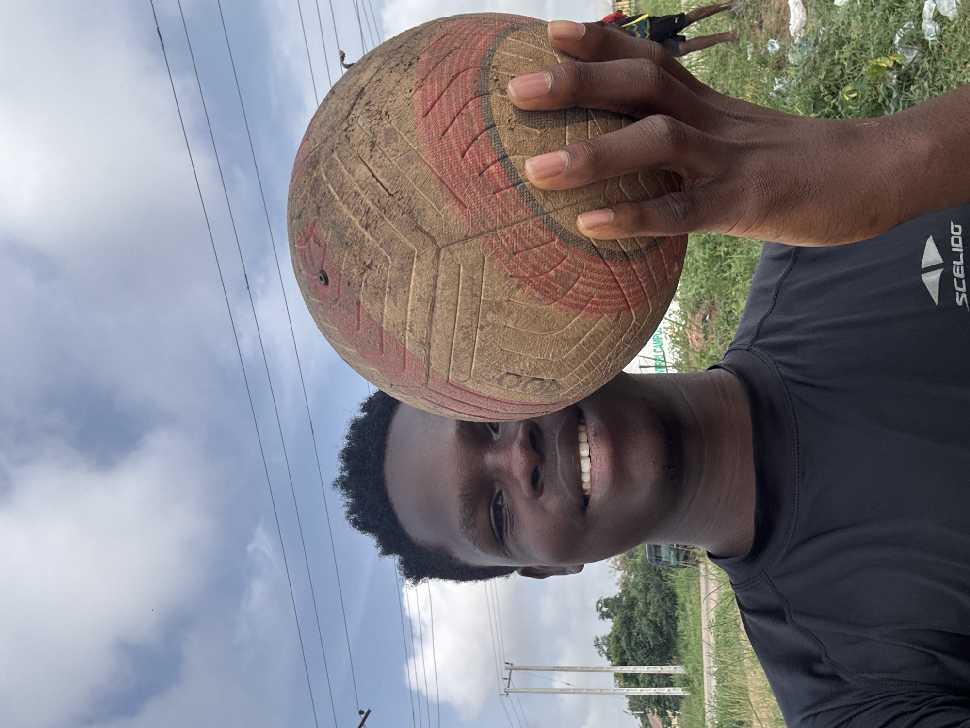 The image is presented vertically, featuring a young man smiling broadly and holding a well-used volleyball with his right hand, positioned above his head. The man has dark skin and short, dark, curly hair. His wide smile reveals white teeth, conveying a sense of happiness or enjoyment.
The volleyball is aged and dirty, showing significant wear, scuffs, and dirt, with faded red and beige/brown panels. The man's fingers are visible, gripping the ball firmly. He wears a dark short-sleeved athletic shirt, likely black, with the white brand name "SCELIDO" and its accompanying logo (two triangles) prominently displayed on his upper chest/shoulder area. A very faint, partial text, possibly "TRALCAMP", is visible on the worn surface of the volleyball.
The background features a partially cloudy sky dominating the upper portion of the frame, traversed by several thin utility wires or power lines. Below, a grassy area with scattered litter and debris, including plastic bags, is visible. Further in the distance, lush green trees and blurry figures of other individuals can be discerned. The overall setting appears to be an informal outdoor space, consistent with the provided context of Ihiagwa, Nigeria, during daylight hours.