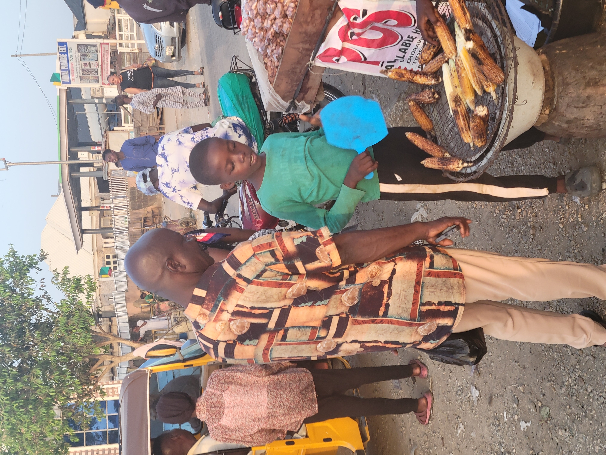 In Madalla, Nigeria, a street vendor is selling grilled corn and possibly other goods from a cart attached to a motorcycle. A boy wearing a green shirt is holding a blue fan near the grilled corn. A man wearing a patterned shirt is standing nearby, appearing to be a customer. Other people are walking in the background. The environment appears to be a street with buildings on both sides. A yellow tricycle is in the foreground. The cart contains corn and what appear to be other goods in a sack. The corn is being cooked on a round grill placed on top of a cylindrical container.