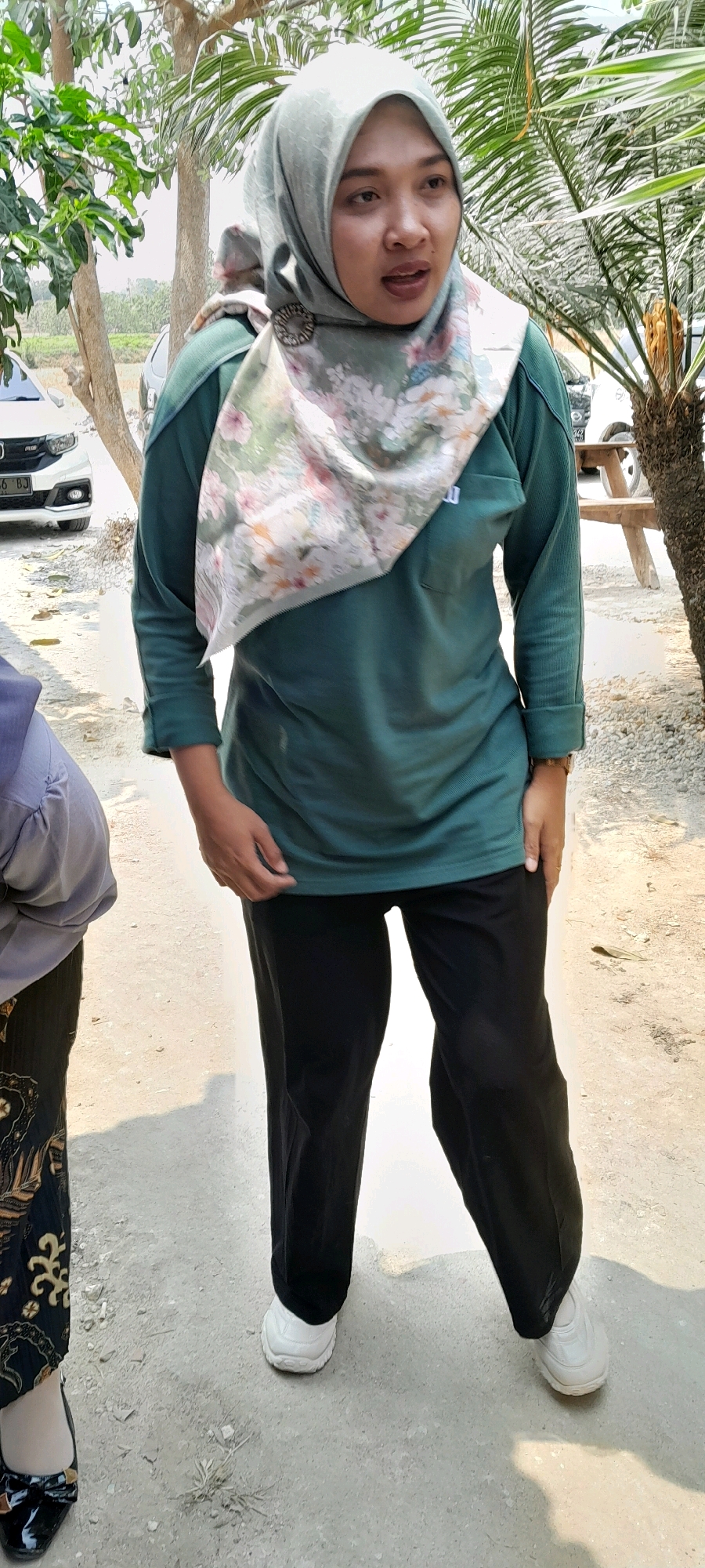 A full-body, outdoor, vertical, medium-shot photograph captures a woman wearing a hijab and standing in a gravelly outdoor area in Sidohasri, Indonesia. The woman is facing slightly to her right, with her mouth open as if speaking. She is wearing a green, long-sleeved top and black pants, along with white sneakers. A light-colored hijab with a floral pattern is draped around her head and shoulders, secured with a circular brooch. To the left of the woman, another person's legs are visible, wearing a patterned skirt and black heels. In the background, a white car is parked, and palm trees and other foliage are present. The lighting suggests it is daytime.