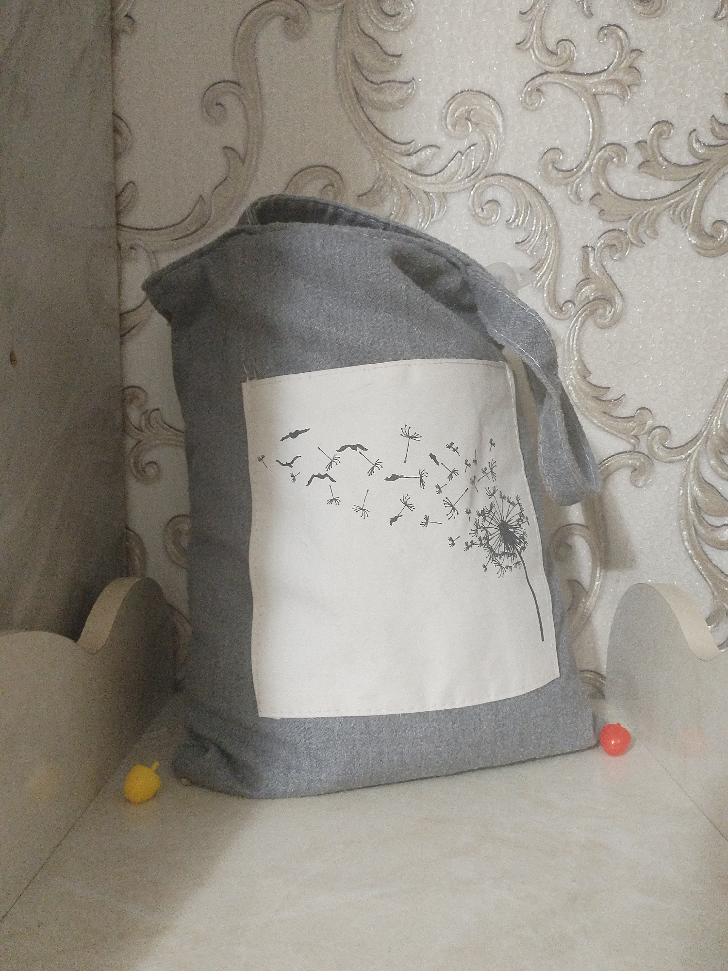 A grey fabric bag with a white rectangular patch on the front featuring a black dandelion silhouette design rests on a shelf against a cream-colored wallpaper with an ornate swirling pattern. Two small, brightly colored objects, one yellow and one red, are placed near the base of the bag on the shelf. The bag has two grey fabric straps. The environment appears to be an indoor setting, possibly a room in a home. There are no people visible in the image. The scene is static and the lighting is soft.