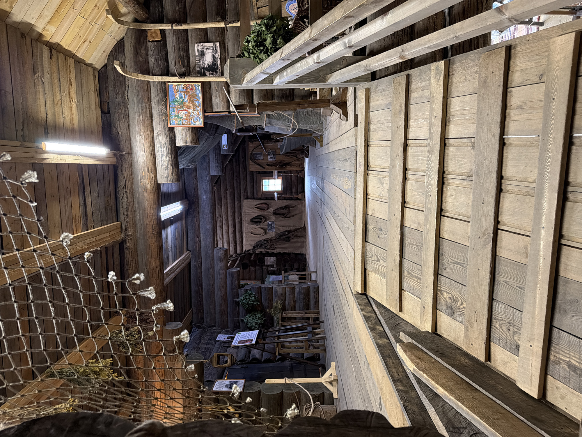 This media file captures an interior view of a rustic wooden structure, likely a museum or a themed exhibit in Imeni Sverdlova, Russia. The scene is dominated by unfinished wooden walls, floors, and ceiling, creating a warm and earthy atmosphere. The lighting is a mix of artificial light from a fluorescent tube fixture and ambient light filtering in from somewhere unseen, suggesting it is daytime.
The composition shows various objects and details that hint at a past or traditional lifestyle, possibly related to fishing or rural life. A prominent feature is a net hanging in the foreground, adding to the rustic, possibly maritime, theme. Further into the structure, there are stacked logs, wooden crates, and what appear to be display boards with information or images. One framed artwork, featuring a colorful, possibly folk art, depiction, hangs on the wooden wall, alongside a black and white photograph of an old building. Small bundles of dried herbs or foliage are also visible, adding to the natural and traditional feel. A small, square window with visible panes is located in a darker section of the structure, admitting a faint light.
There are no people visible in the frame, so no activity or event involving humans is taking place. The overall impression is one of a thoughtfully curated space designed to evoke a specific historical or cultural context, likely a local history or ethnographic exhibit. The wooden textures and the displayed objects contribute to a sense of authenticity and groundedness. No discernible text is clearly legible in the provided image.