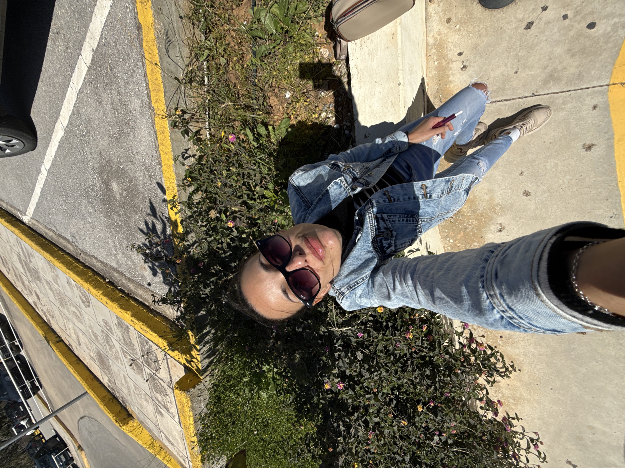 A woman lies in some shrubbery near a sidewalk, next to a parking lot in Mouzouras, Greece. She wears ripped jeans, a black top, and a denim jacket. She is also wearing sunglasses. There is a tan-colored purse nearby. A car is parked in the parking lot.