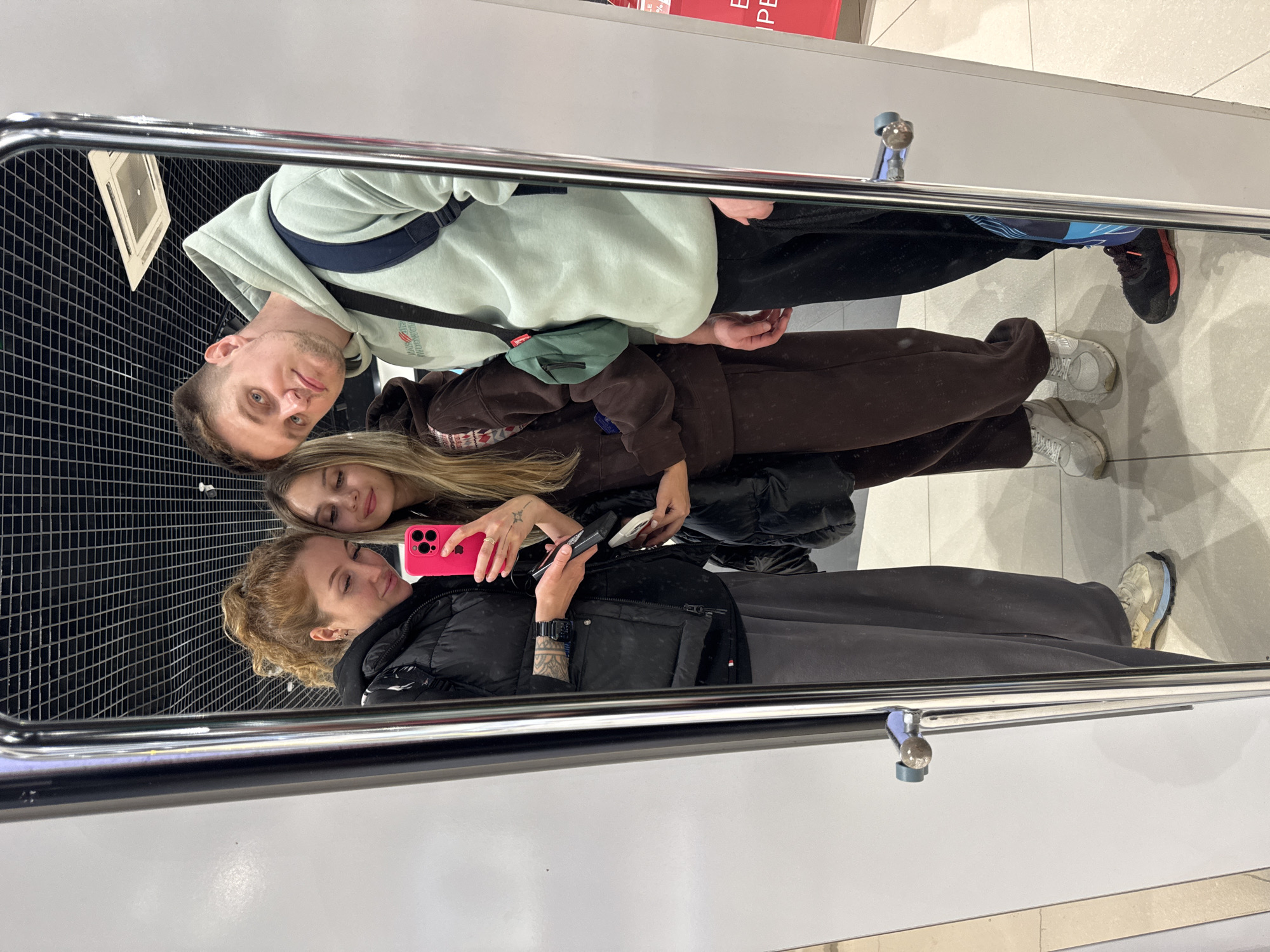 Three individuals are reflected in a mirror. One person wears a light green hoodie and black pants. Another individual wears a brown sweatsuit. The third individual is wearing a black vest and dark grey pants. The individual in the middle is holding a pink smartphone. The setting seems to be indoors, possibly a store or shopping mall. Sochi, Russia, is not directly visible or discernible from the image.