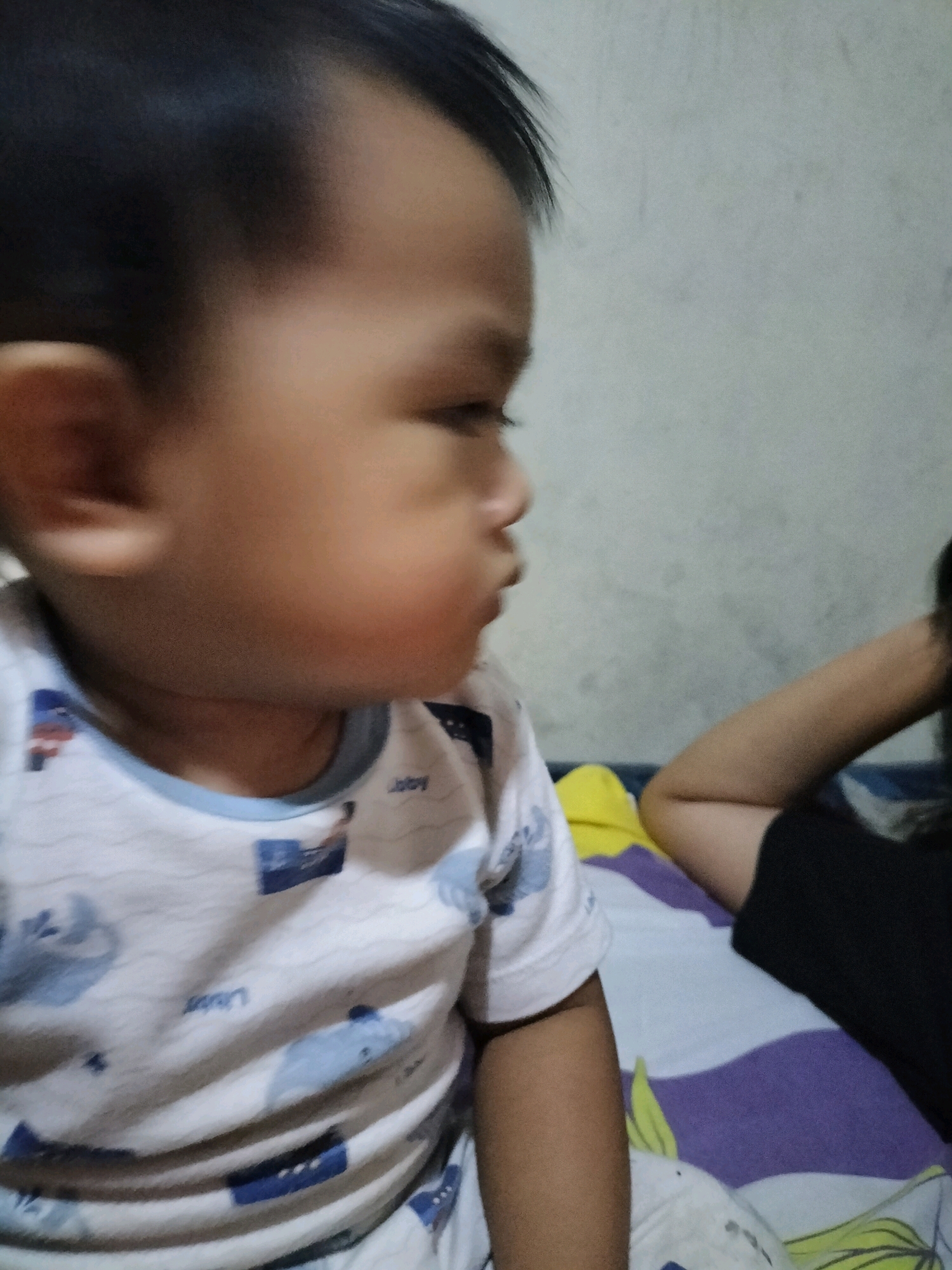 A close-up, side-profile, medium shot captures a toddler facing right, resting on a bed. The toddler's hair is dark and short, and they wear a white t-shirt with a blue trim and a repeating pattern of blue whales and boats. Their left ear is visible. To the right, an adult's arm and shoulder are visible, wearing a black garment. The bed is covered with a striped comforter in purple, white, and yellow, with a subtle floral pattern. The background is a plain, lightly textured wall. The image appears to be taken indoors, possibly in a residential setting in Tasikmadu, Indonesia.