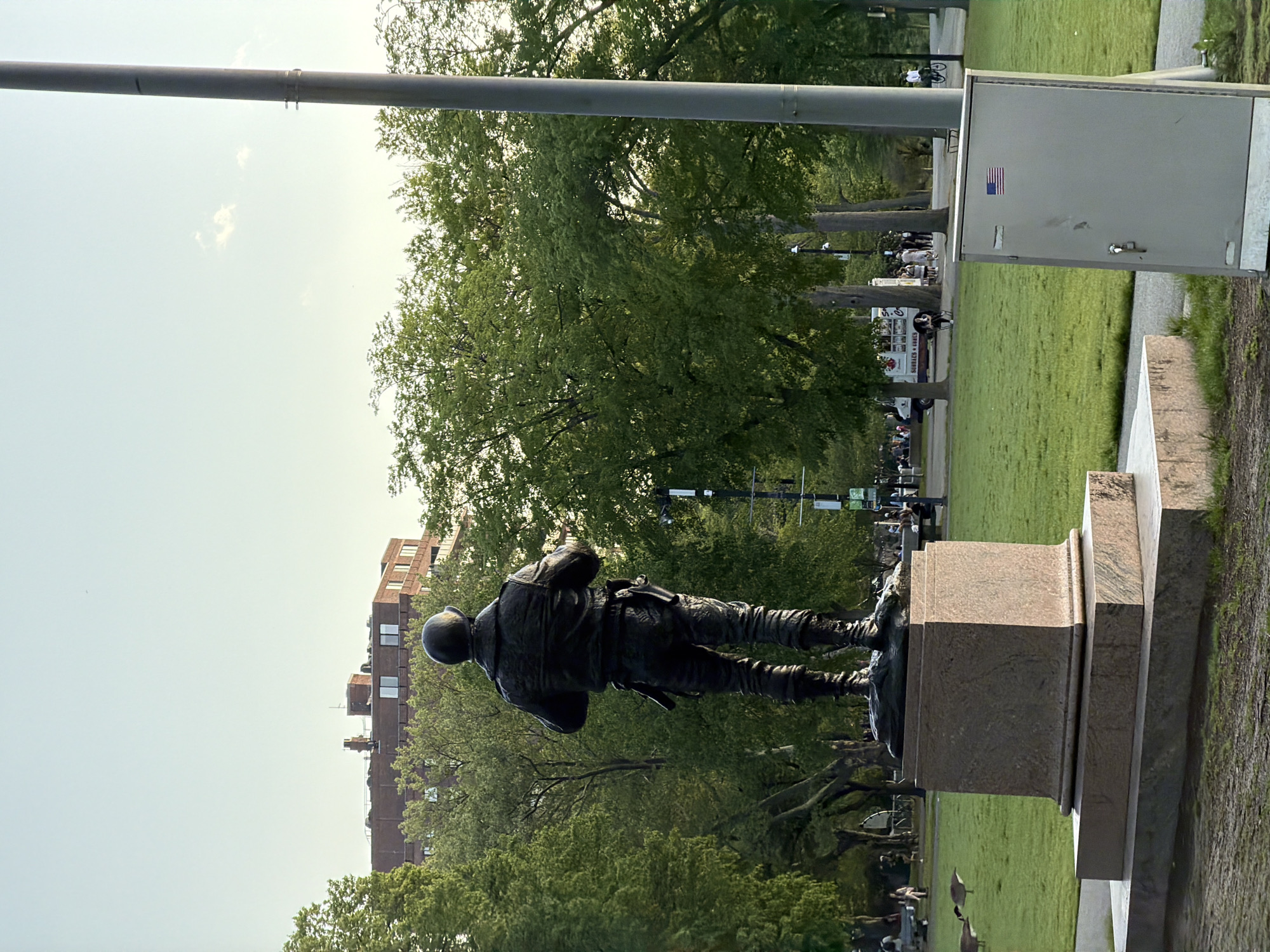 The image depicts a bronze statue of a soldier standing on a granite pedestal. The statue is located within the Boston Public Garden. The soldier is wearing military attire, including a helmet, jacket, and trousers. In the background, there are green trees, grass, and a tall brick building. A metal pole and an electrical box with an American flag sticker are also visible in the scene. People are walking around in the park.