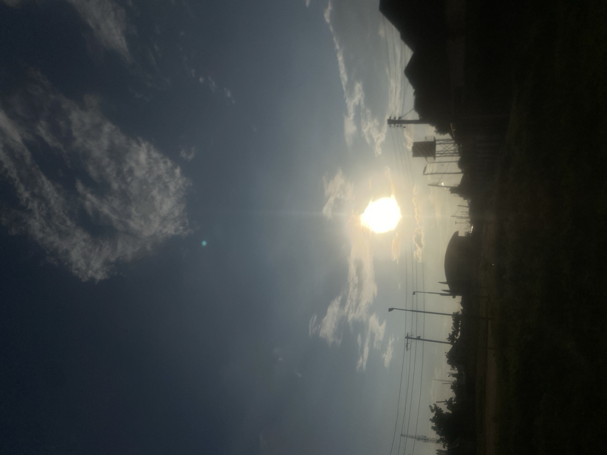 The image shows a silhouette of buildings and infrastructure against a bright sky with the sun setting or rising. The location is identified as Barden Yana, Nigeria. In the foreground, there is a dark, out-of-focus area that appears to be a field or ground. Behind this, a row of silhouetted structures includes buildings, utility poles with wires, and a water tower. The sun is a prominent, bright orb in the sky, casting rays and creating a lens flare effect. There are scattered clouds in the sky. The overall impression is of an urban or semi-urban landscape at twilight.
