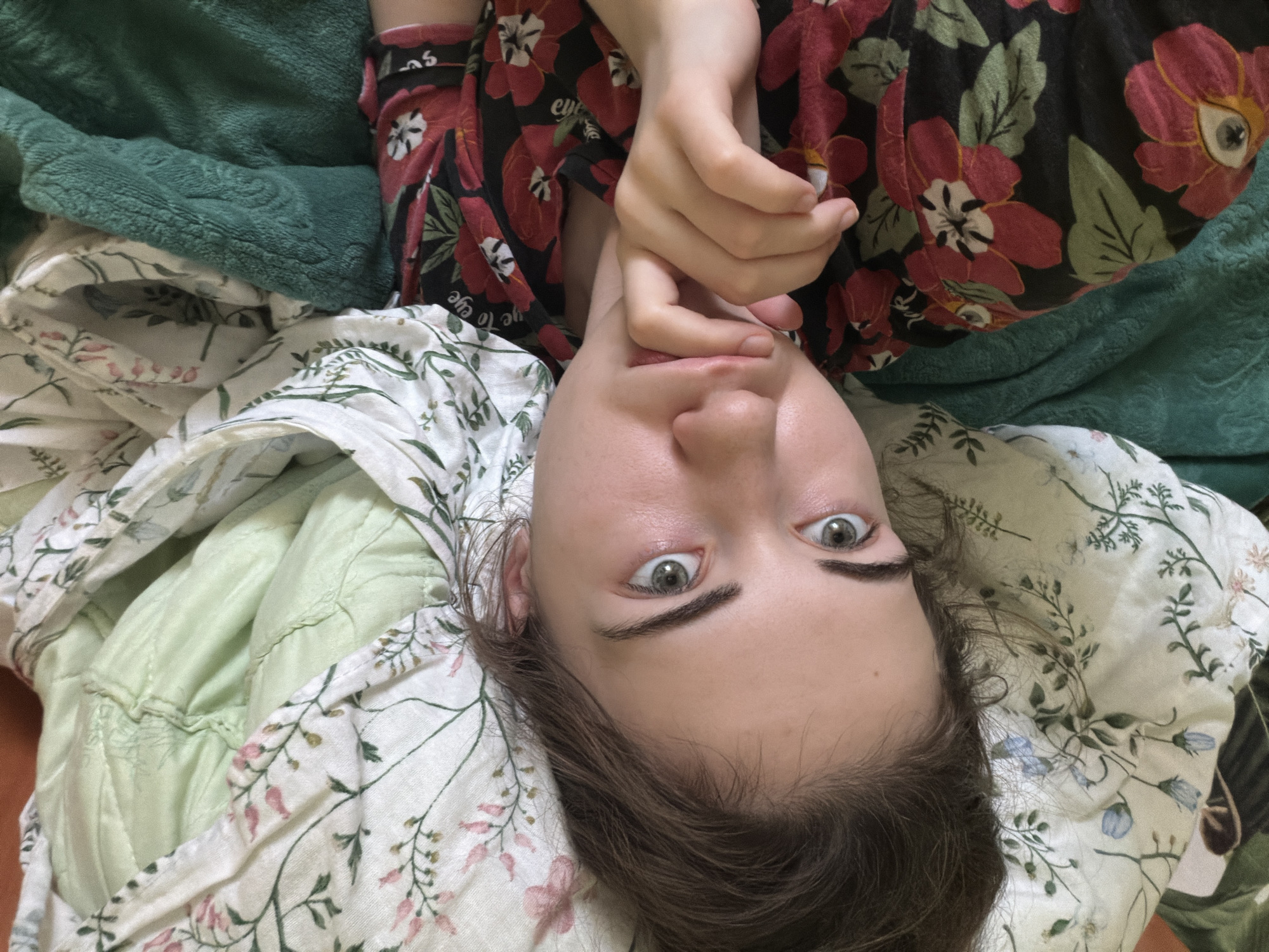 A young woman with light skin and brown hair is lying on a bed covered with floral-patterned sheets and a green blanket. She is looking at the camera, with her hand resting on her chin. Her eyes are green and she has a neutral expression on her face. The room appears to be a bedroom, with soft lighting and a peaceful atmosphere. The woman is wearing a dark floral-patterned shirt. The image is taken from a high angle, looking down at the woman. It is not possible to determine the location in the city of Zavodoukovsk, Russia from the provided image.