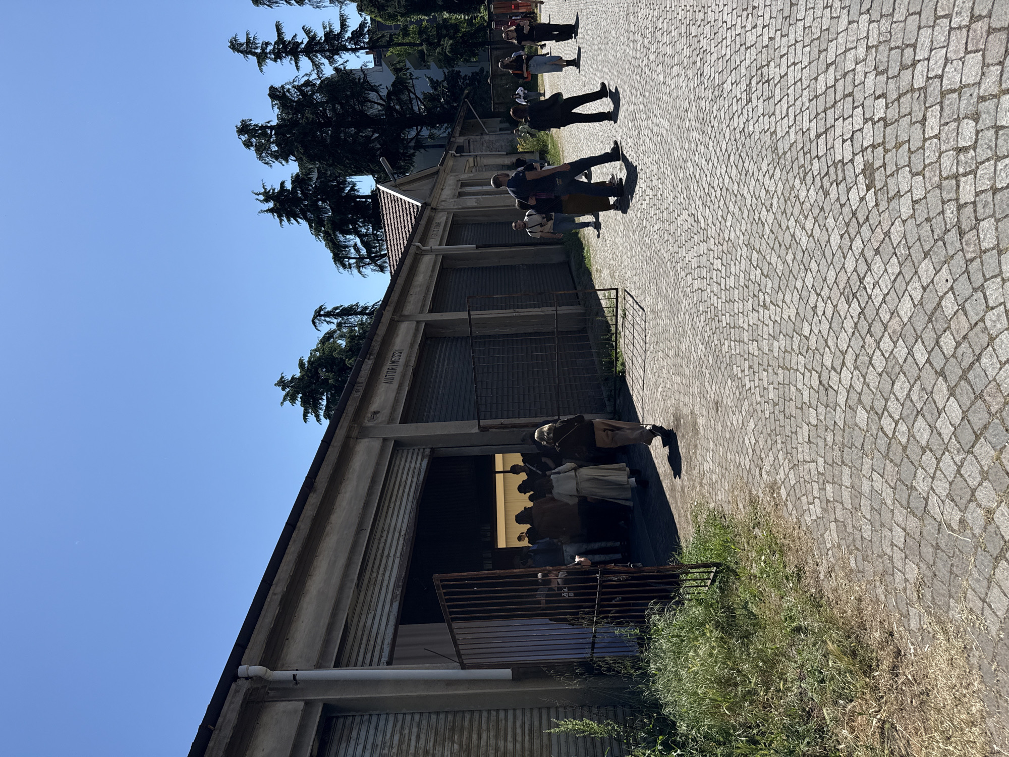 The image captures a street scene in Milan, Italy, featuring a long, low, concrete building with multiple bays, some closed with metal roll-up doors and one open. The building appears to be old, with "AUTORIMESSA" inscribed above the bays. People are walking past the building on a cobblestone walkway. Some individuals appear to be entering or exiting the open bay, where the interior is dimly lit. Tall evergreen trees are visible behind the building against a clear blue sky. Vegetation grows along the base of the building and adjacent to the walkway. A metal railing is present near the open bay. The scene suggests an urban, historical context.