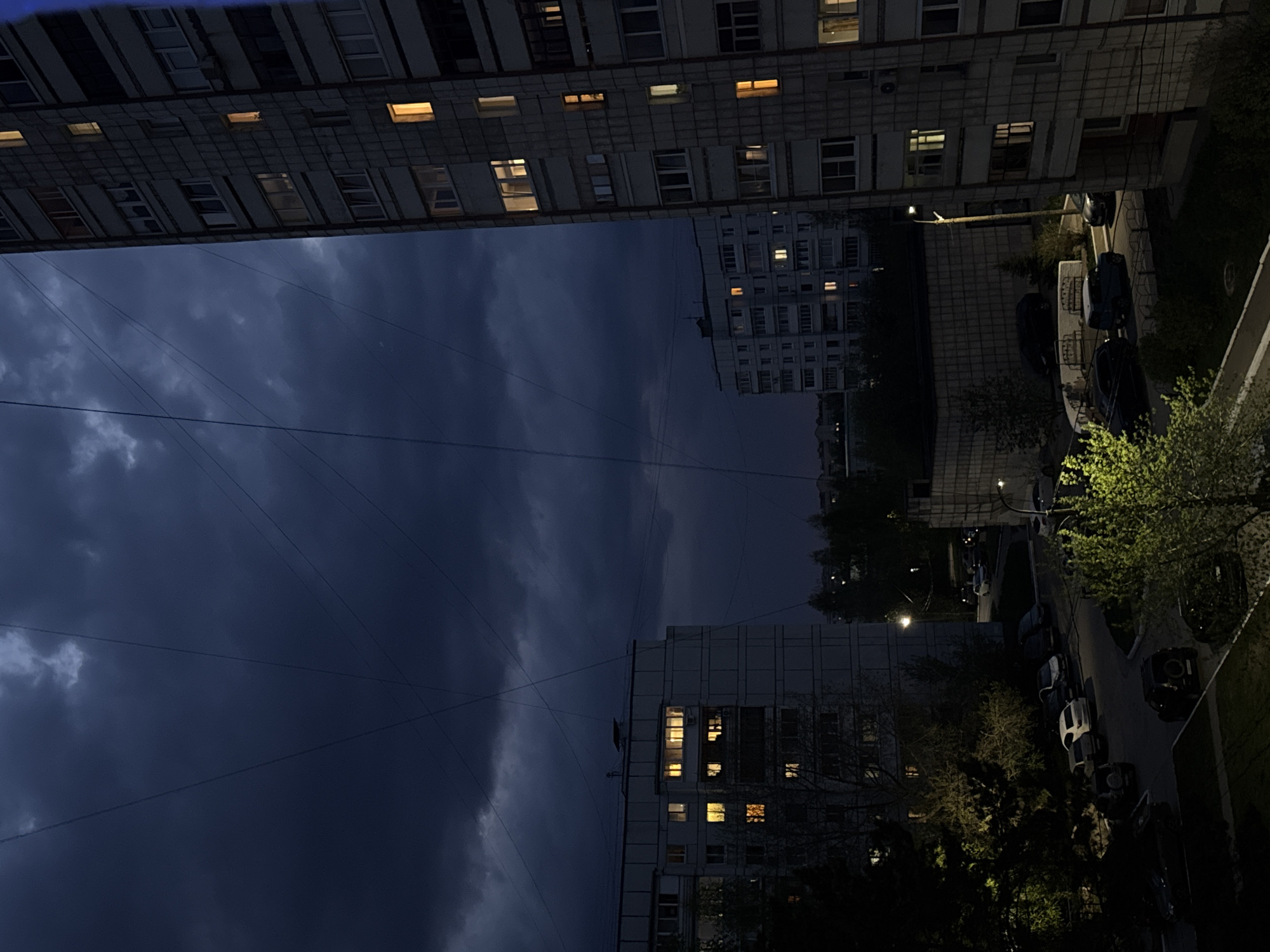 View from an elevated position looking out over a streetscape in the city of Chaikovsky, Russia at night. The scene includes several multi-story buildings, some with lit windows. Utility wires traverse the sky. The street is lined with parked cars and trees. The sky is dark with cloudy formations.